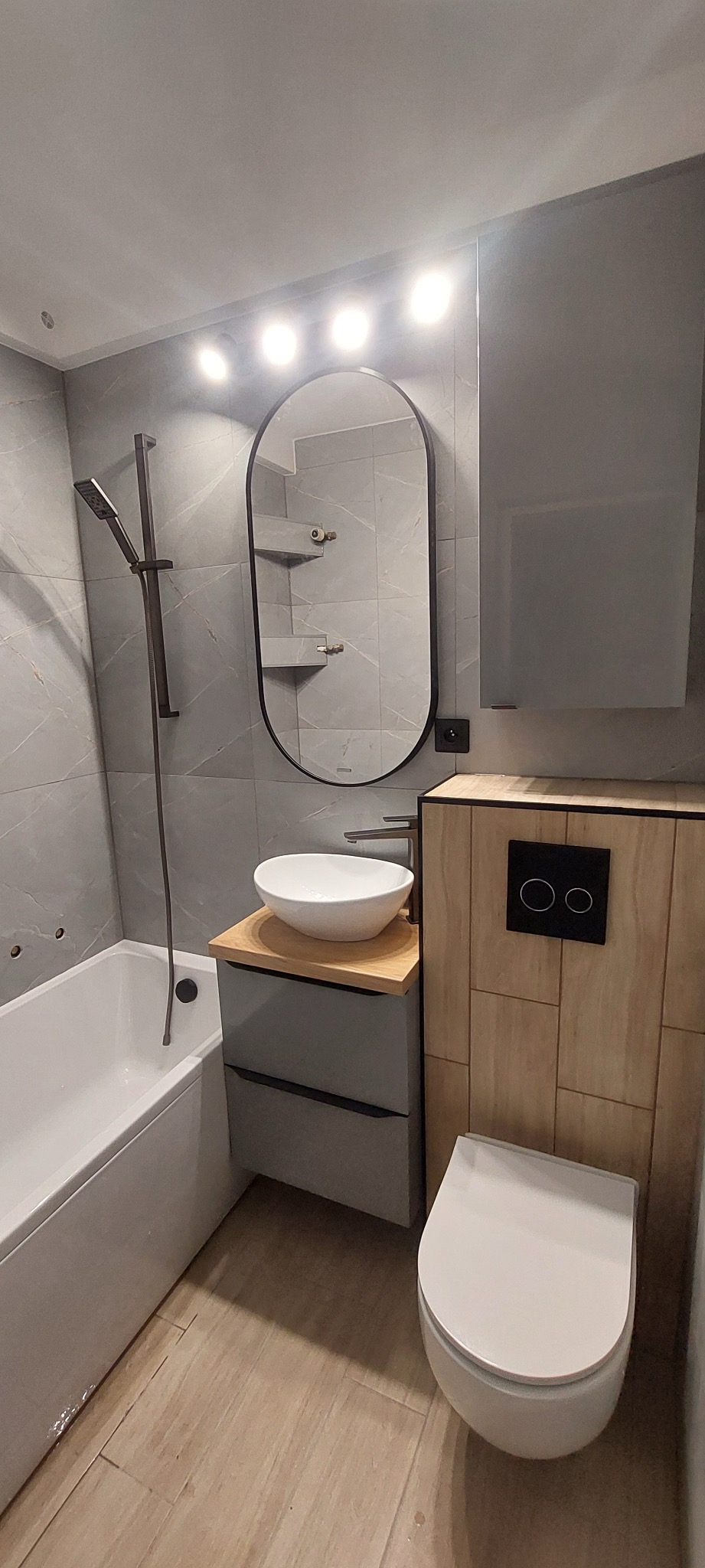 Nowoczesna łazienka w odcieniach szarości i drewna z owalnym lustrem podświetlonym punktowymi lampami, wiszącą toaletą z drewnianą obudową i umywalką nablatową na szarej szafce z drewnianym blatem.