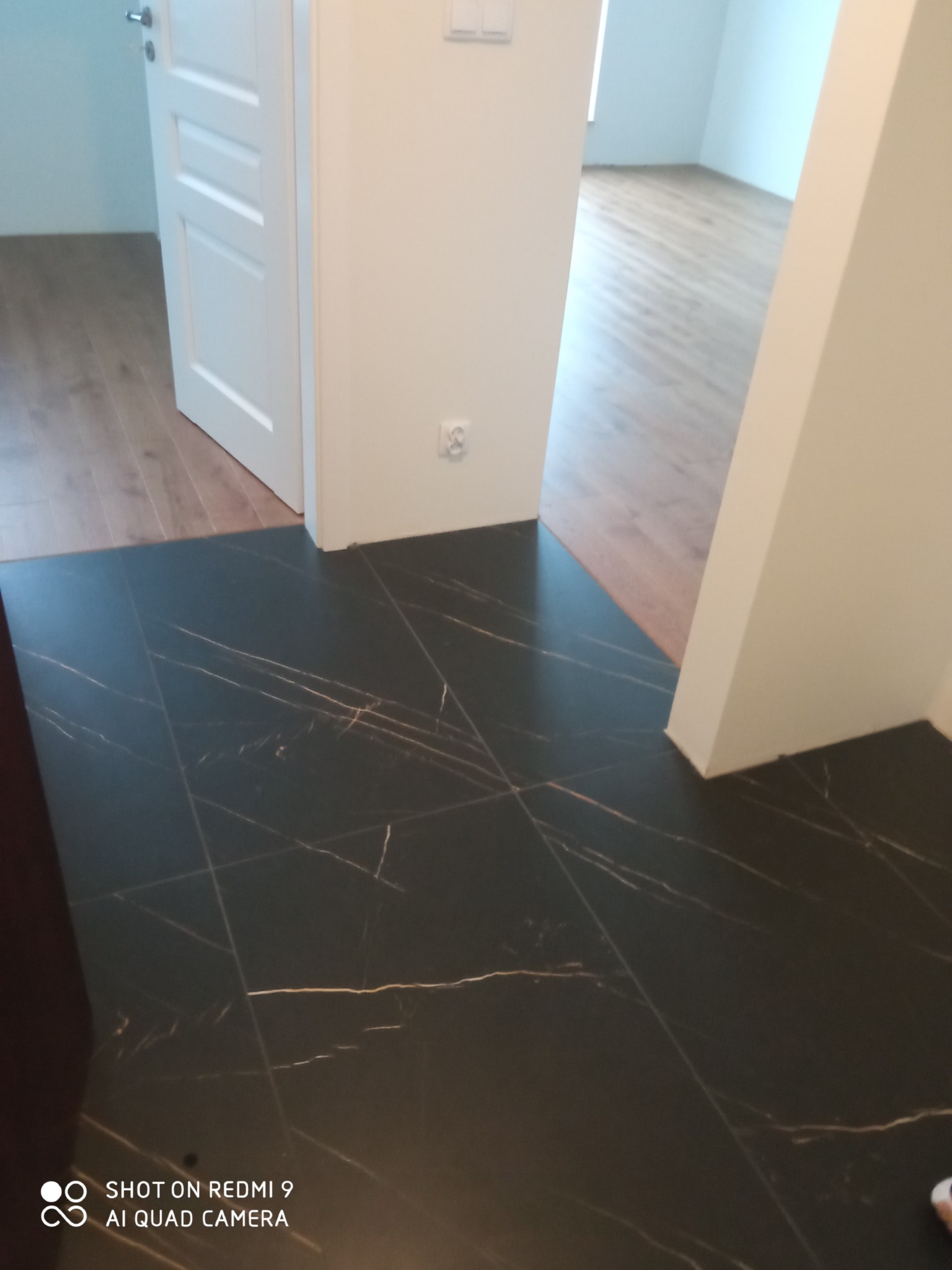 Wykończony fragment podłogi z czarnych płytek z marmurkowym wzorem, widok na odnowione pomieszczenie z drewnianą podłogą przez otwarte białe drzwi.