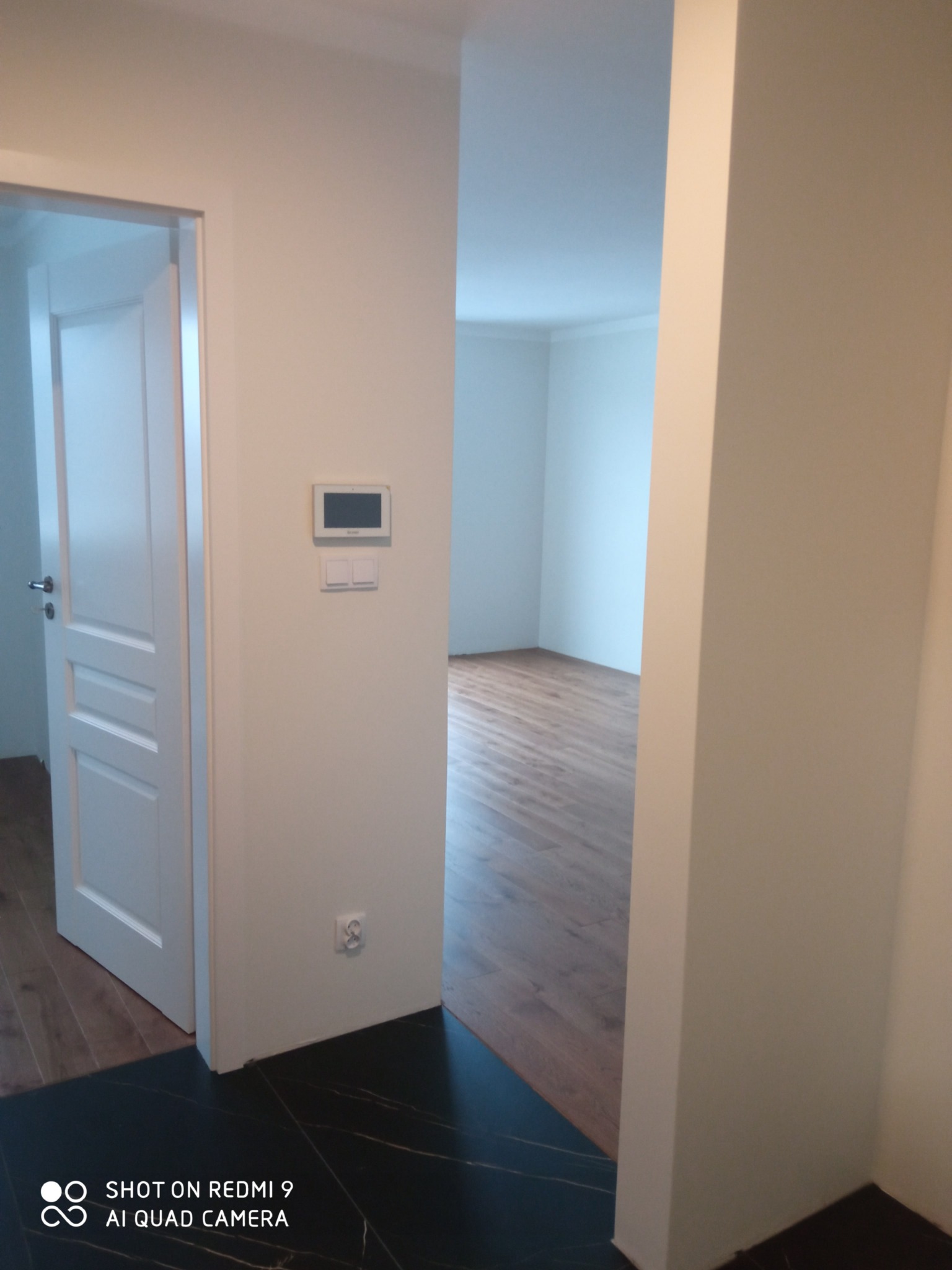 Wnętrze mieszkania po remoncie: białe ściany, ciemna marmurowa podłoga w przedpokoju przechodząca w drewnianą podłogę w pokoju, widoczny panel sterowania na ścianie i otwarte białe drzwi.