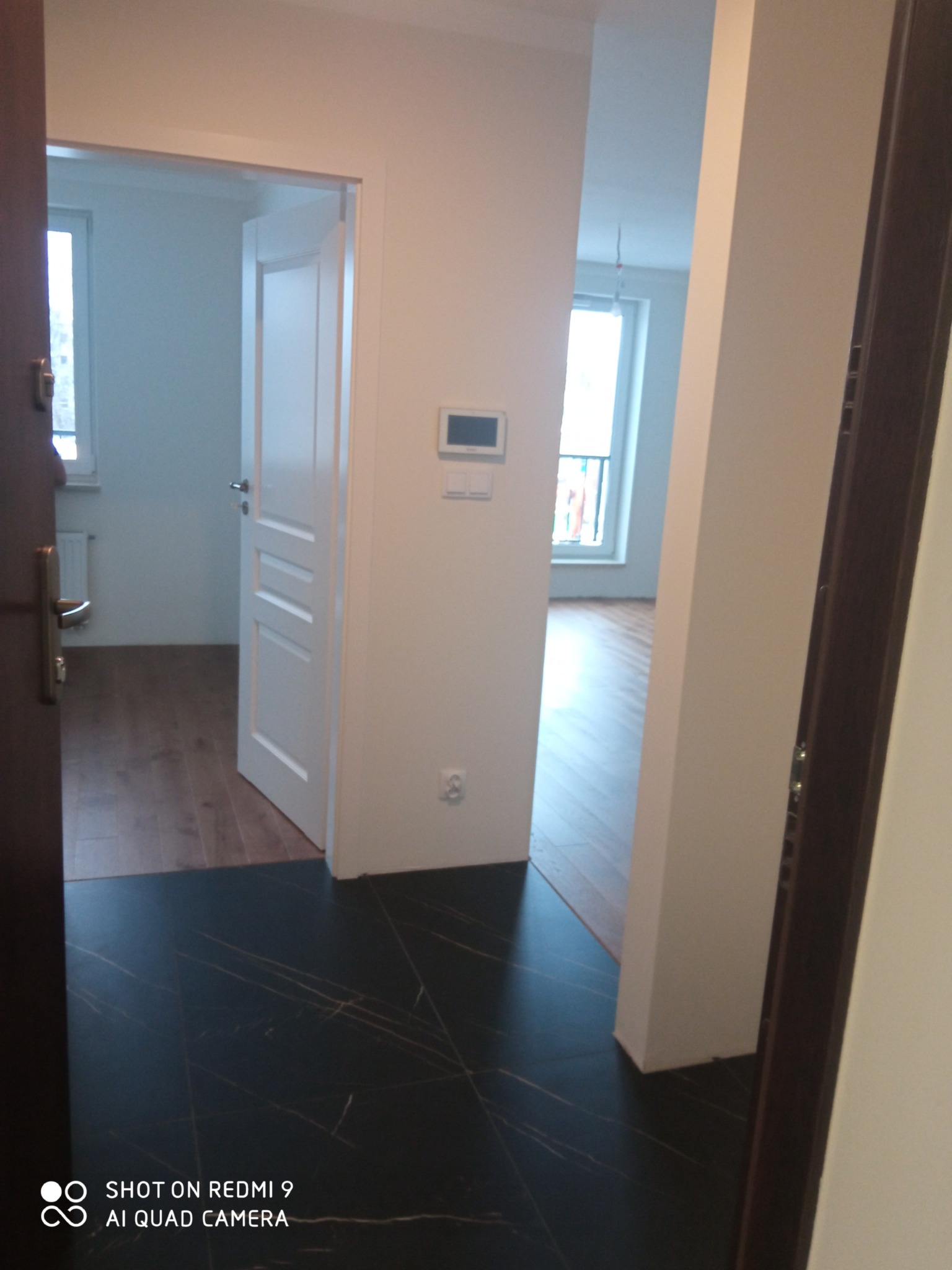 Wnętrze mieszkania po remoncie: widok z korytarza na dwa pokoje z drewnianą podłogą, białe ściany i okna, czarna marmurowa podłoga w korytarzu.