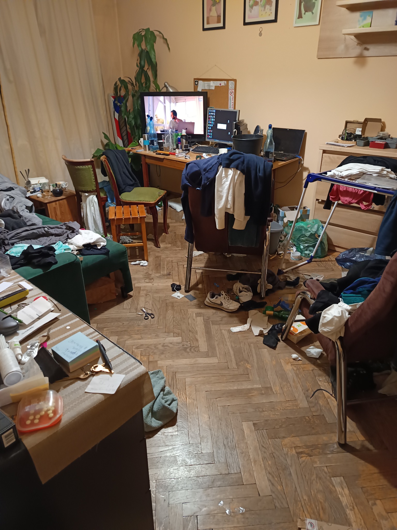 Bardzo zagracony pokój z biurkiem, komputerem, suszarką na pranie i porozrzucanymi ubraniami na podłodze i meblach. Na biurku widoczne liczne przedmioty i butelka wody.