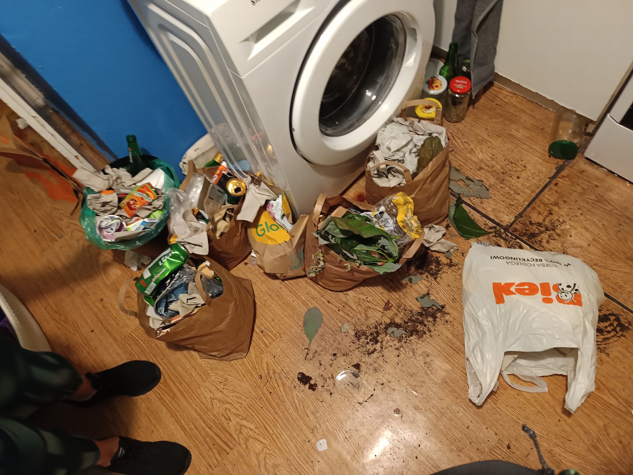 Prowizoryczne worki na śmieci z odpadami budowlanymi i resztkami roślin w pobliżu pralki w trakcie sprzątania po remoncie, z widocznymi butelkami i rozsypaną ziemią na podłodze.