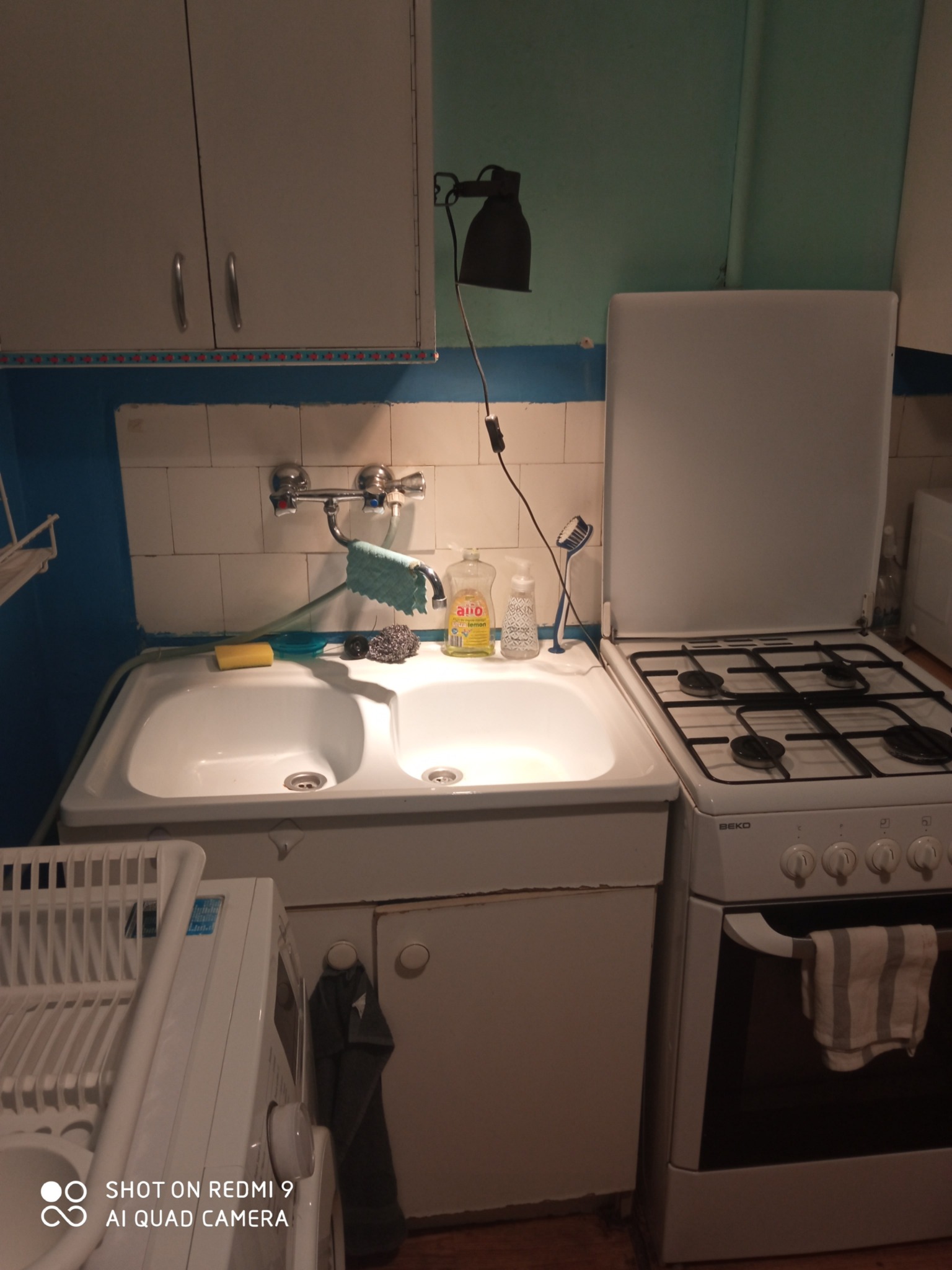 Kuchnia w bloku: biały zlewozmywak dwukomorowy, bateria, płyn do naczyń, gąbka, zmywak; obok kuchenka gazowa Beko, pralka i suszarka na naczynia.