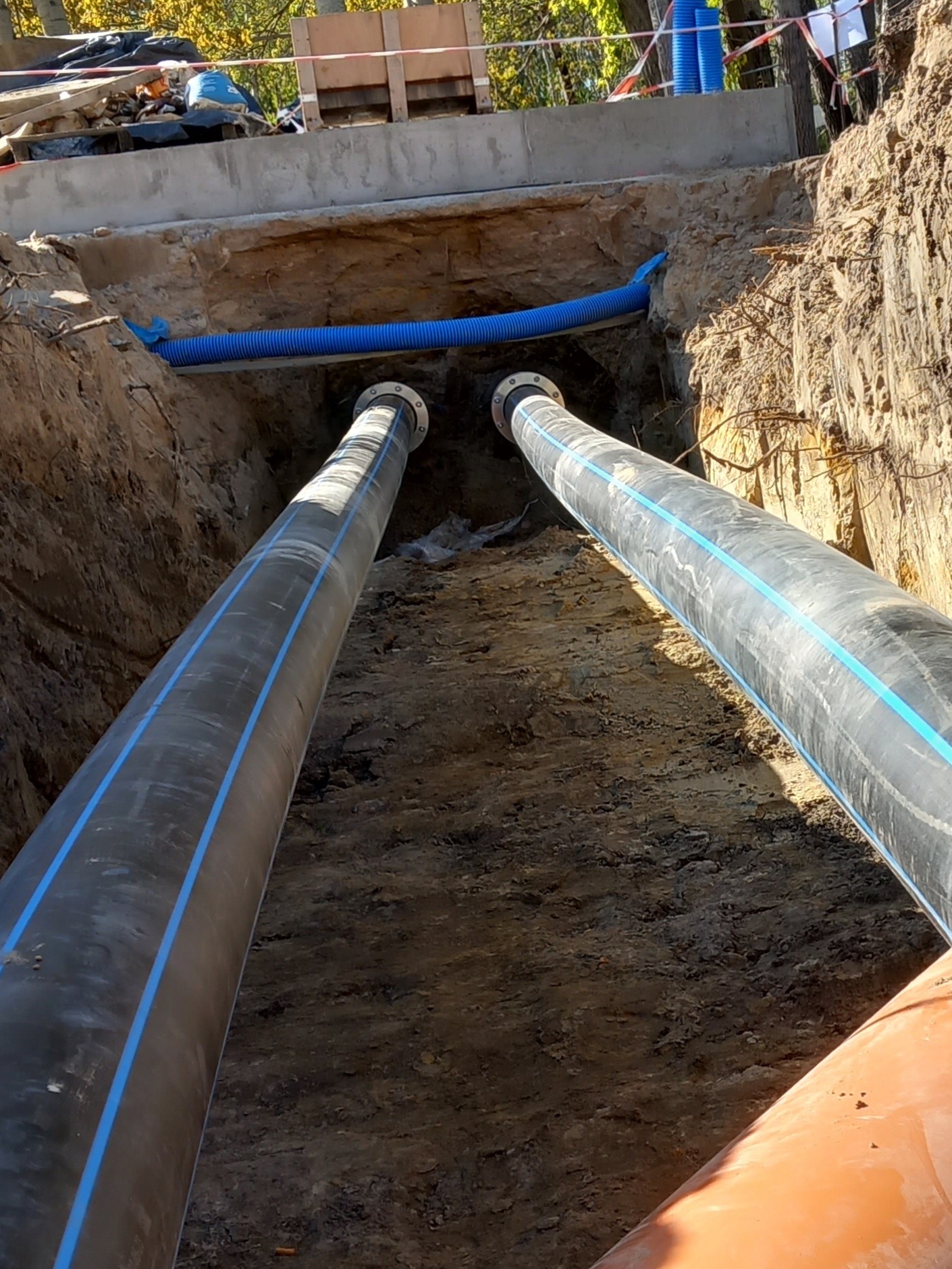Dwie równoległe rury z niebieskimi paskami ułożone w wykopie ziemnym z widocznymi fragmentami niebieskiej rury karbowanej i betonowej konstrukcji powyżej.