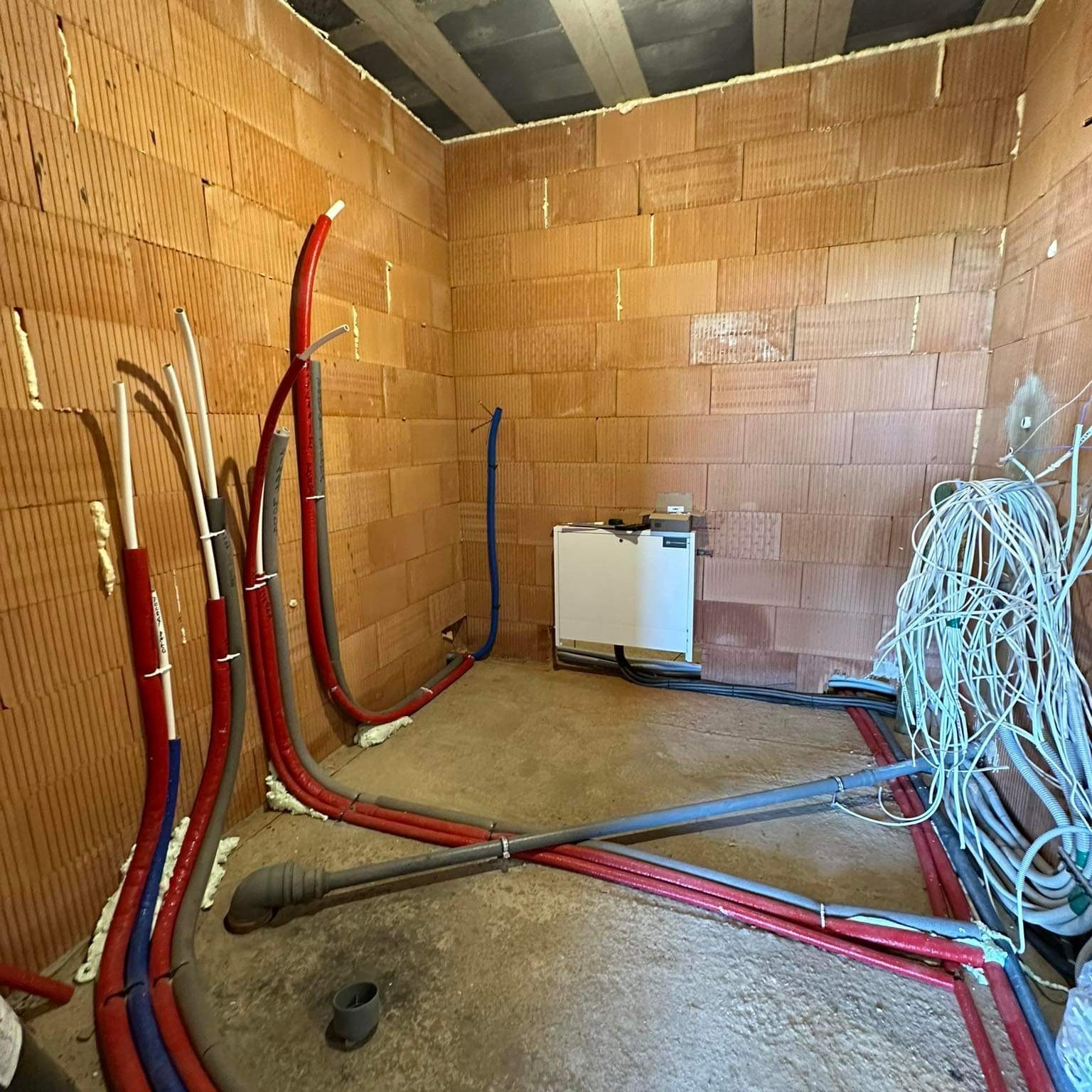 Surowy stan pomieszczenia z instalacjami hydraulicznymi i grzewczymi w trakcie montażu: rury w izolacji czerwonej i niebieskiej, biały rozdzielacz na ścianie z cegły, plątanina przewodów...