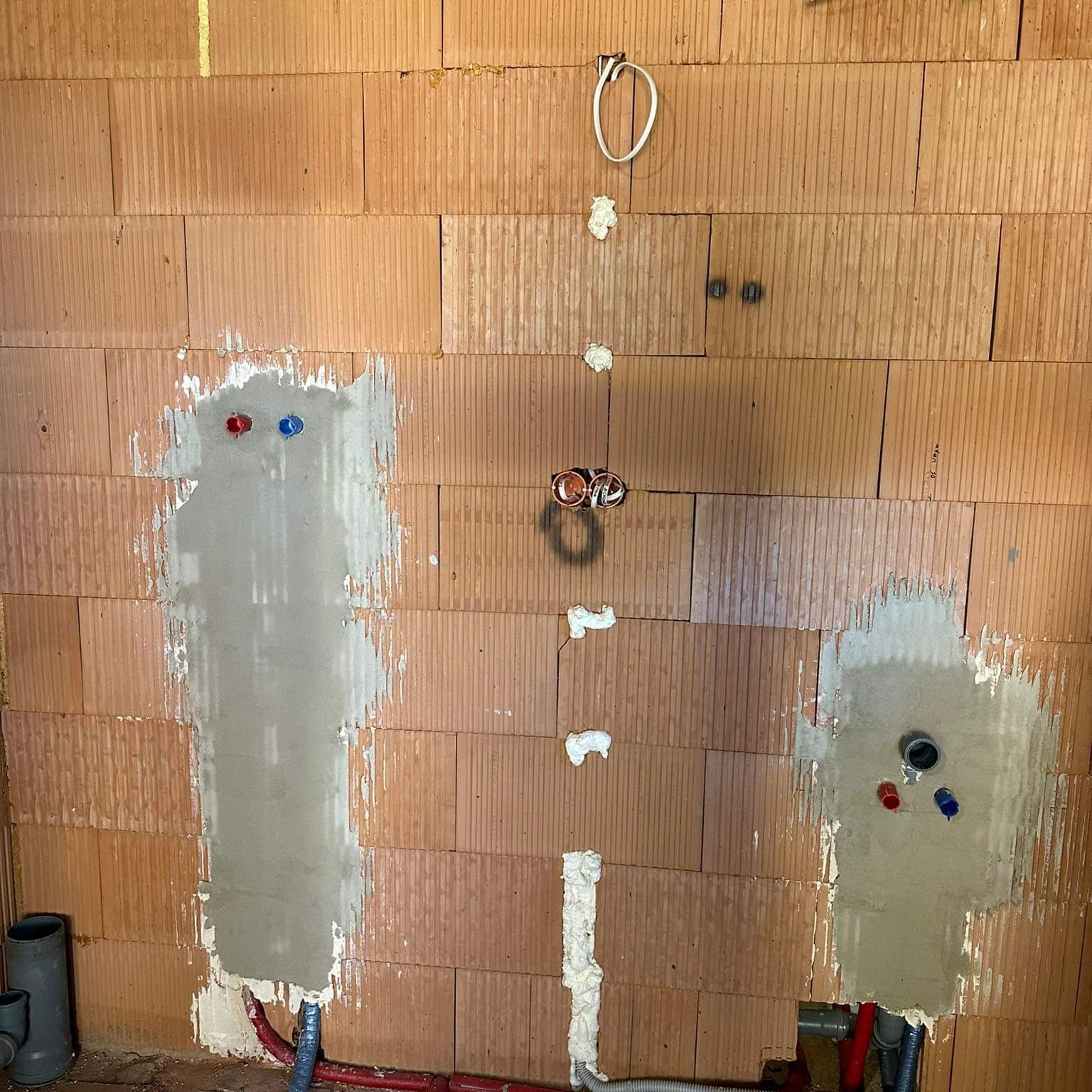 Ściana z pustaków ceramicznych z wykuciami pod instalacje wodne i elektryczne, częściowo zamurowane. Widoczne rury PEX w kolorze czerwonym i niebieskim oraz przewody elektryczne. Wykucia...