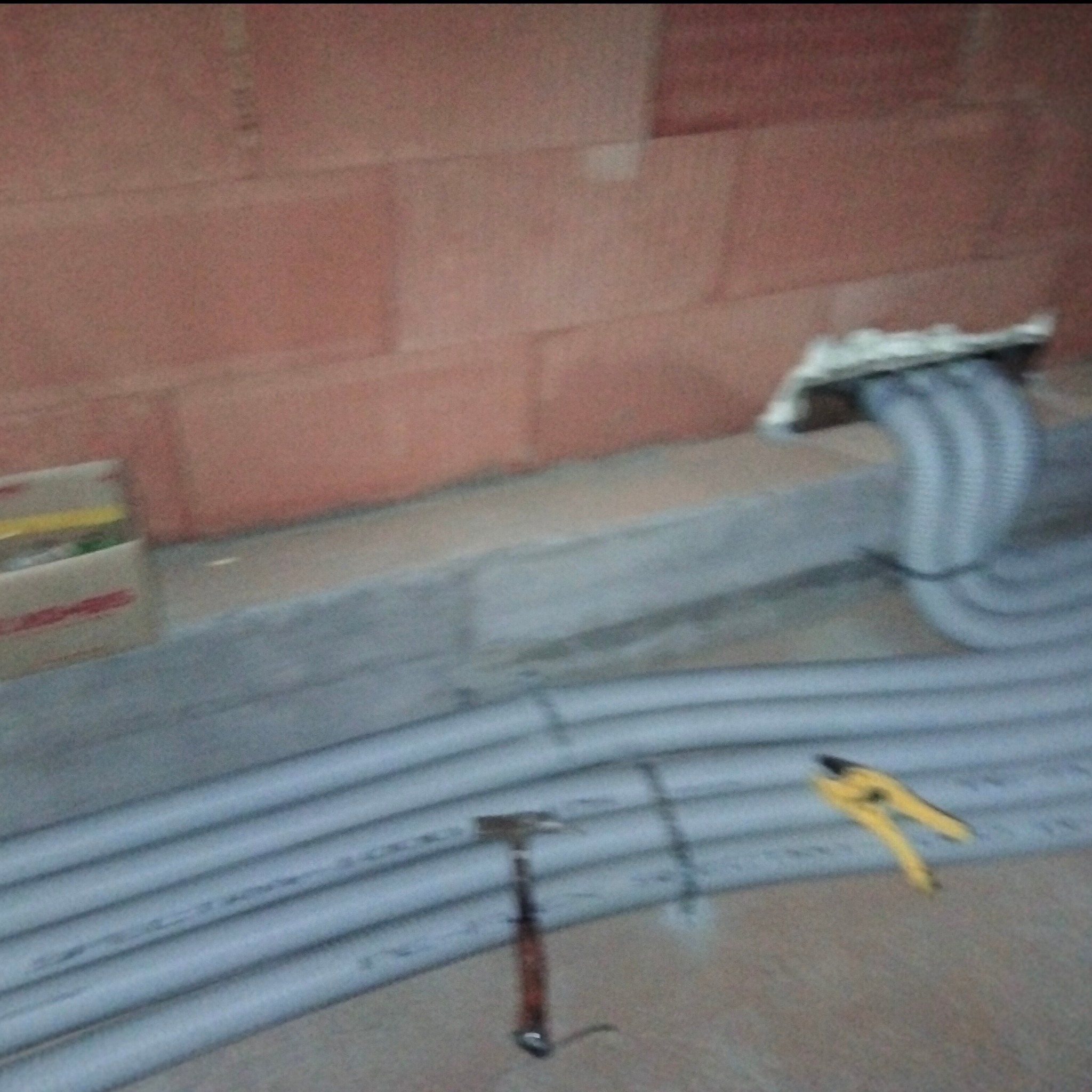 Szare rury instalacyjne ułożone w bruździe w ścianie z cegły, widoczne narzędzia: młotek i obcinaczki do rur.