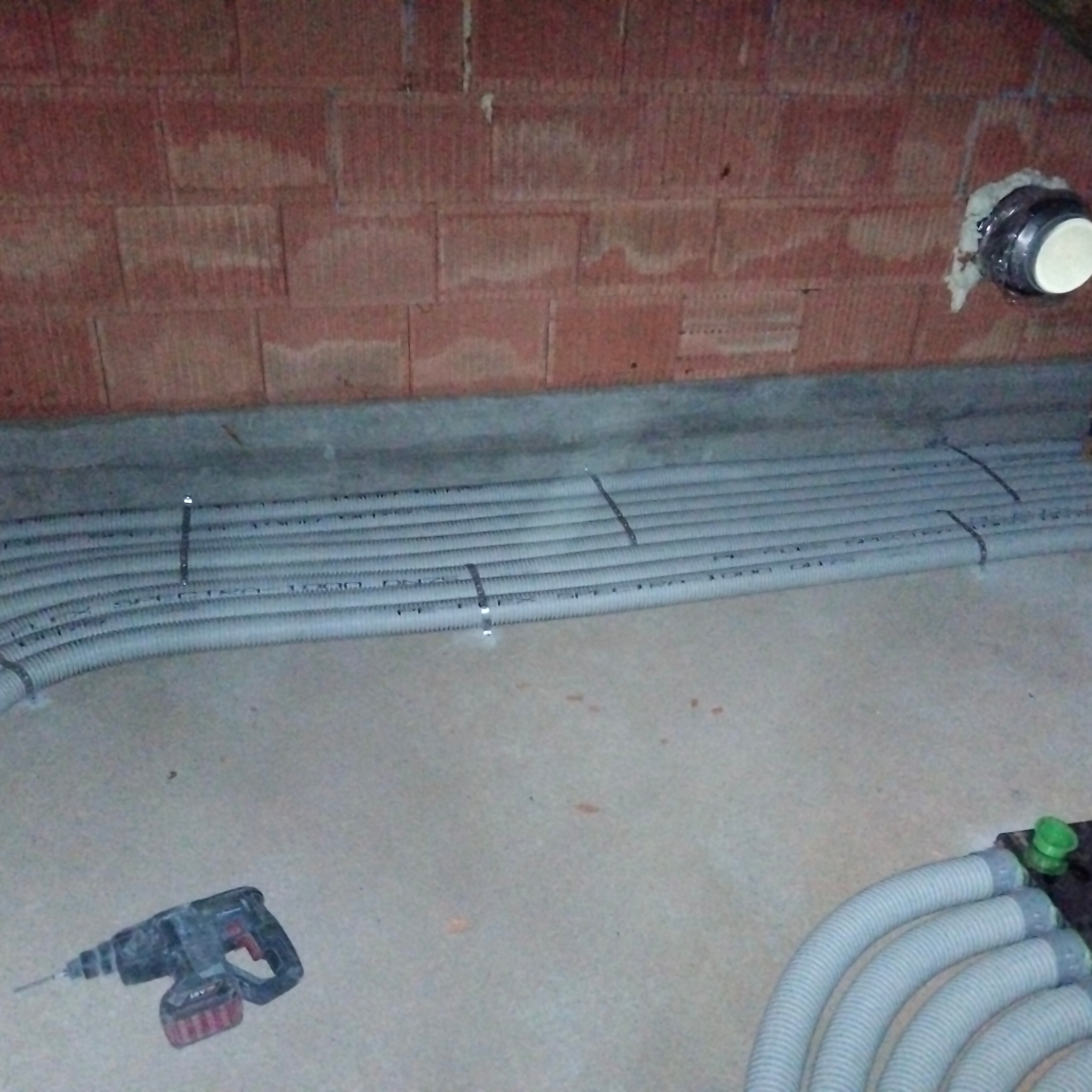 Szare rury instalacyjne ułożone równolegle na podłodze w surowym stanie, przymocowane do podłoża metalowymi klamrami, widoczna ściana z cegły dziurawki oraz wiertarka akumulatorowa w tle.