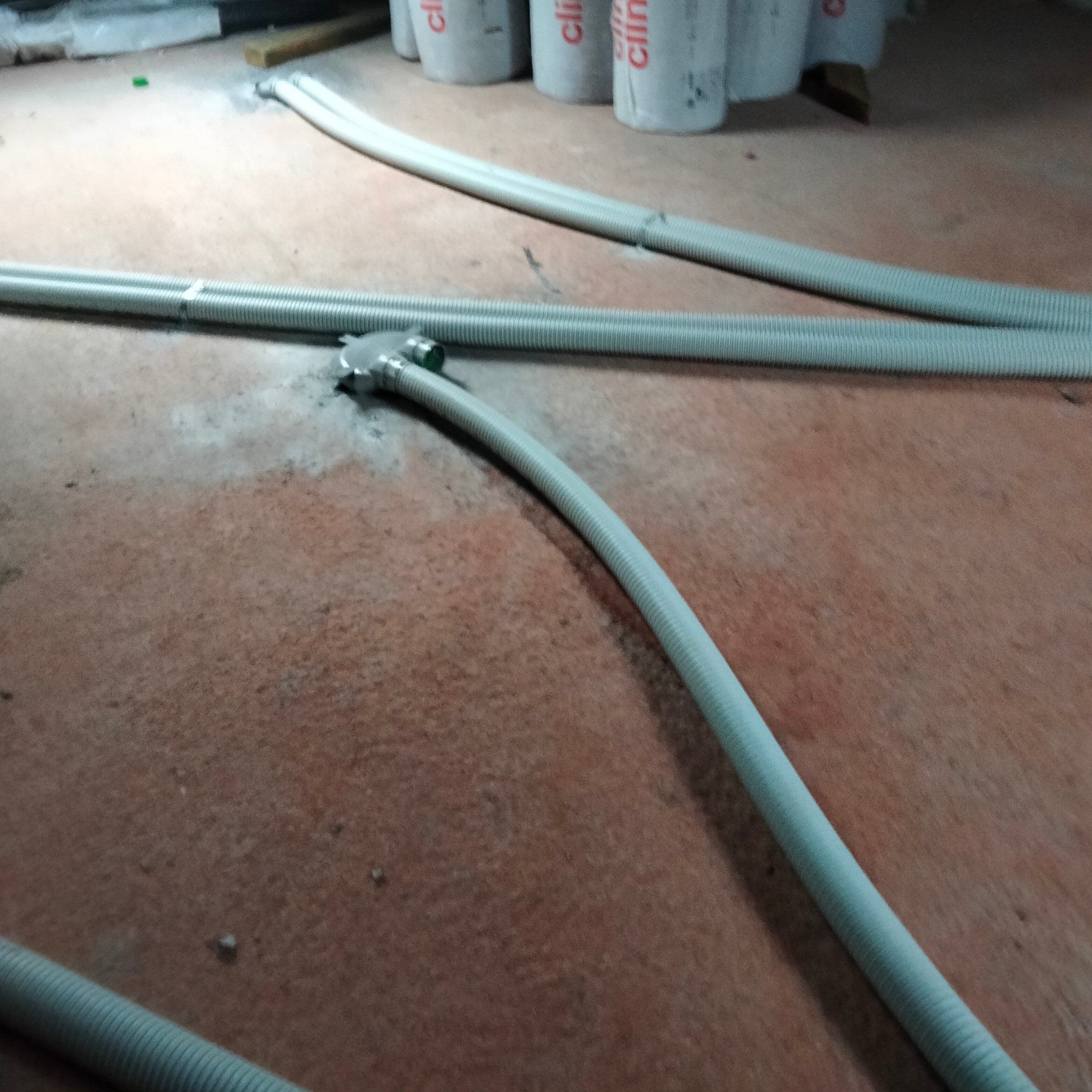 Fragment instalacji ogrzewania podłogowego z szarymi rurami karbowanymi na tle brązowej posadzki, widoczne białe pojemniki z napisem 'clima'.