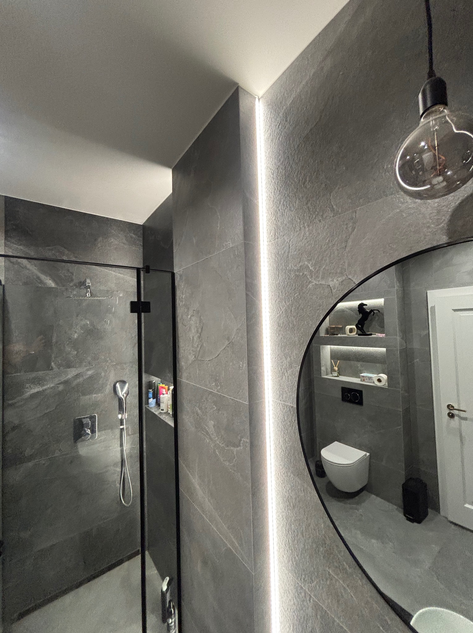 Nowoczesna łazienka z szarymi płytkami imitującymi kamień, kabiną prysznicową typu walk-in i okrągłym lustrem odbijającym toaletę. Oświetlenie LED podkreśla pionowe linie.