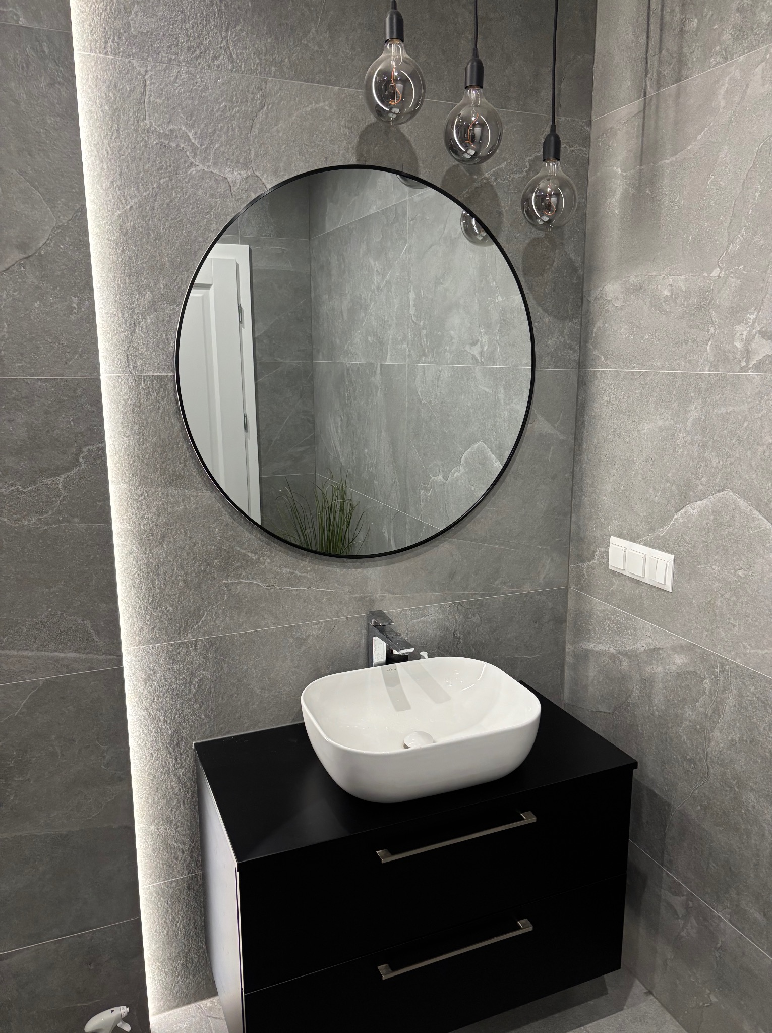 Szara łazienka z okrągłym lustrem, umywalką nablatową na czarnej szafce i trzema żarówkami dekoracyjnymi. Płytki imitujące kamień z podświetleniem LED.