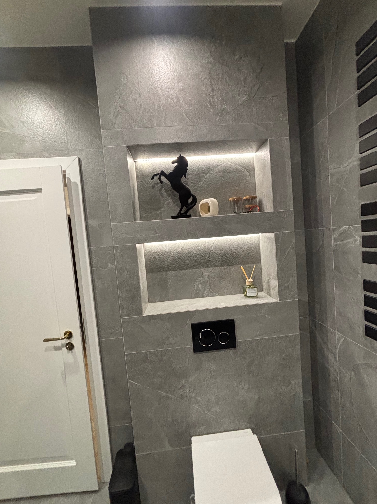 Stylowa łazienka z szarymi płytkami imitującymi kamień, podświetlane wnęki z dekoracjami, biała armatura i drzwi z złotą klamką. Minimalistyczny design.