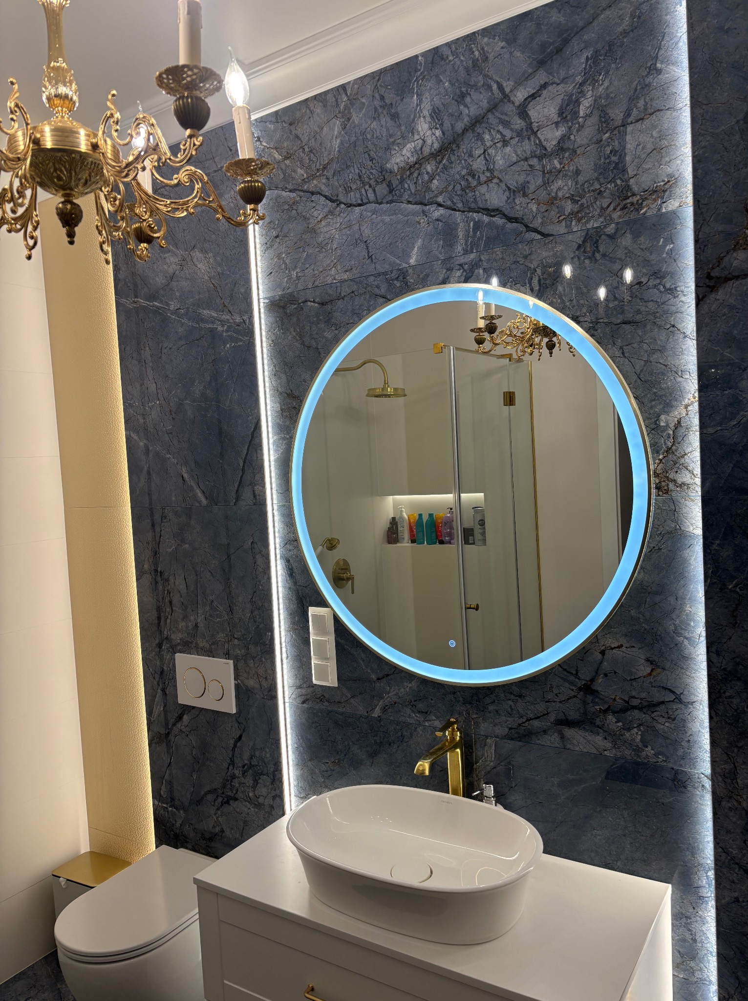Elegancka łazienka z marmurową ścianą, okrągłym lustrem z podświetleniem LED, złotą armaturą i ozdobnym żyrandolem. Widoczna umywalka nablatowa i toaleta.