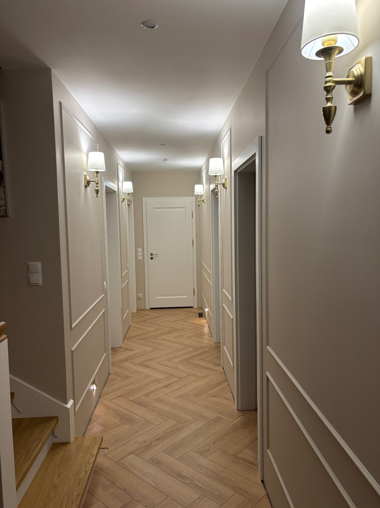Korytarz w odcieniach beżu z parkietem ułożonym w jodełkę, oświetlony złotymi kinkietami. Ściany zdobione sztukaterią, białe drzwi. Widok z perspektywy schodów.