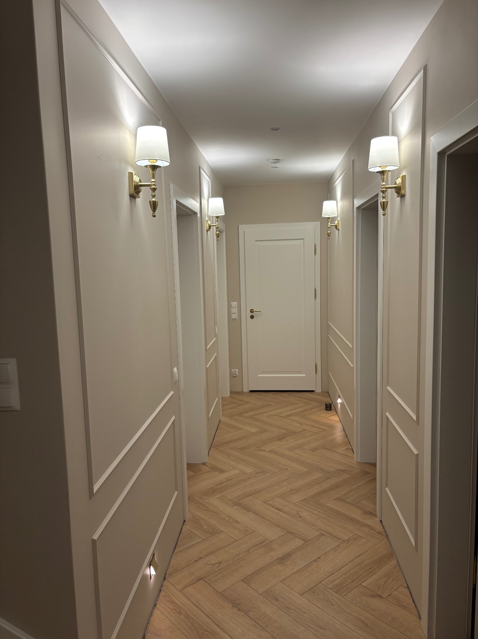 Korytarz po metamorfozie: parkiet w jodełkę, kremowe ściany z dekoracyjnymi listwami, złote kinkiety. Nowoczesne wykończenie wnętrza z dbałością o detale.