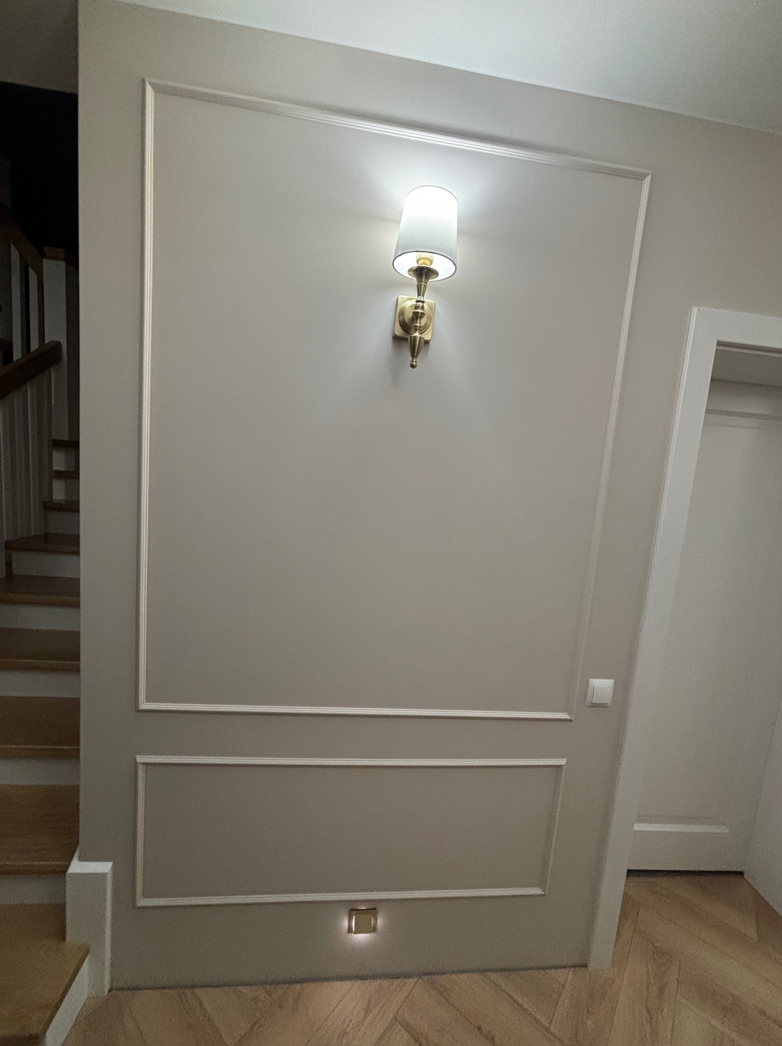 Ściana w odcieniach beżu z dekoracyjnymi listwami i kinkietem w stylu retro. Parkiet ułożony w jodełkę. Widoczne schody i fragment drzwi.