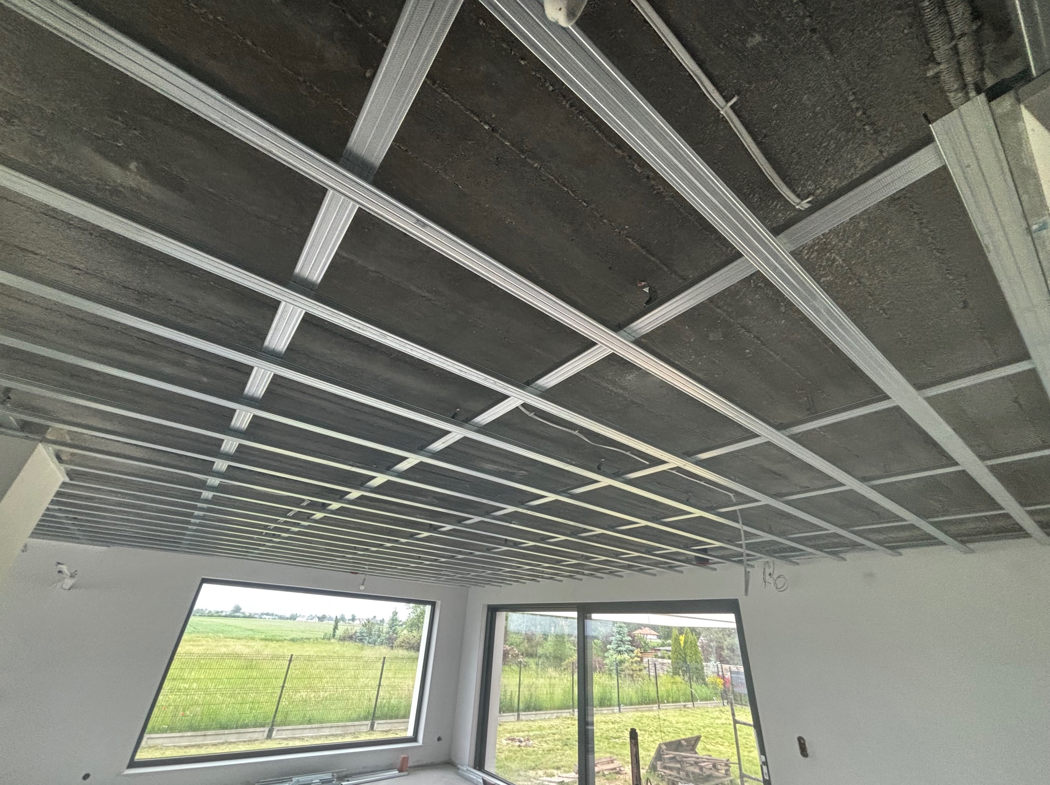 Metalowa konstrukcja sufitu podwieszanego z płyt karton-gips, widok z dołu, surowy betonowy strop, okna z widokiem na zieleń w tle.