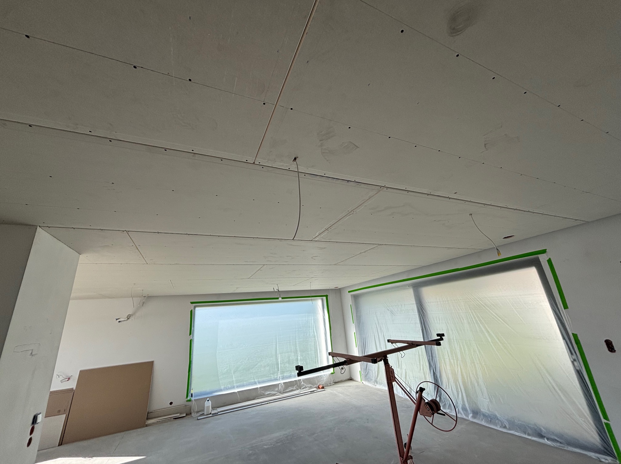 Sufit z płyt gipsowo-kartonowych w trakcie montażu, widoczne łączenia i wkręty. Okna zabezpieczone folią malarską, na podłodze narzędzia i resztki materiałów.