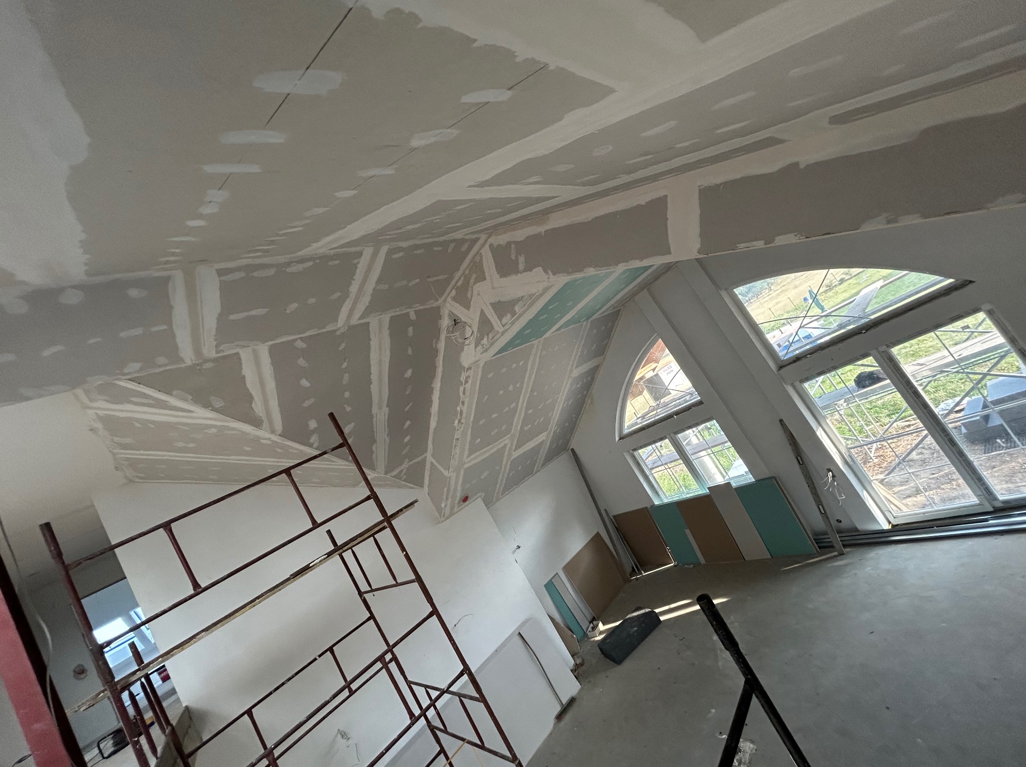 Skośny sufit z płyt kartonowo-gipsowych w trakcie montażu, widoczne łączenia i szpachlowanie. W tle okna i rusztowanie. Jasne wnętrze w stanie surowym.