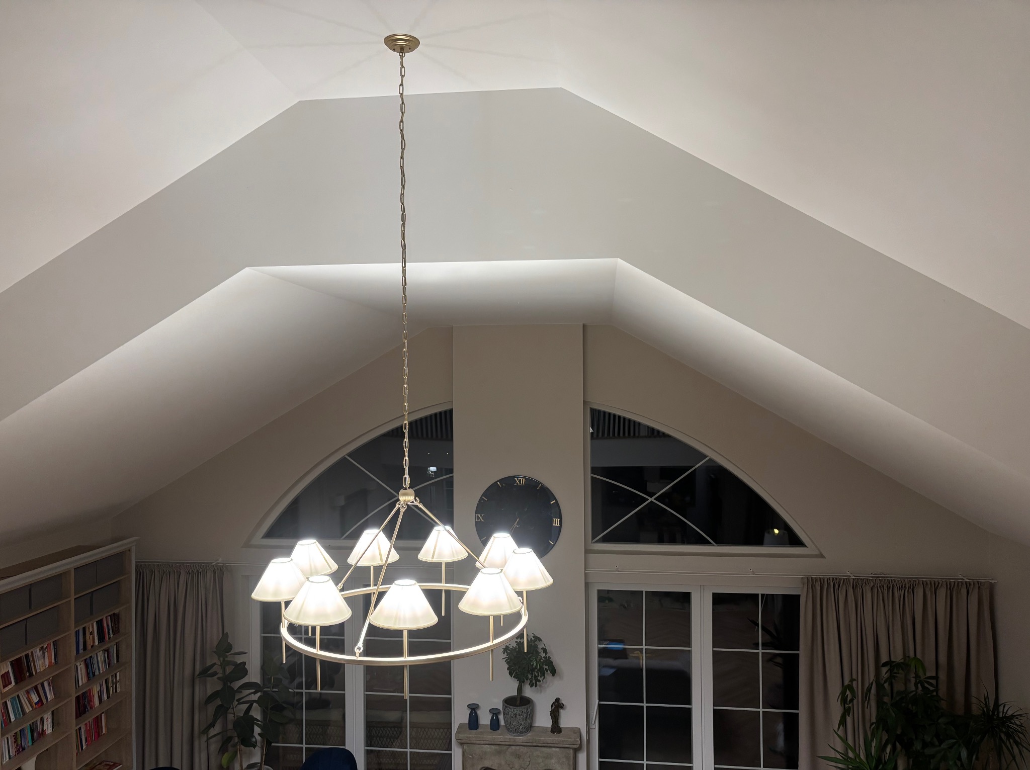 Jasne wnętrze z wysokim sufitem i oknami łukowymi, oświetlone przez wiszący żyrandol. Widoczna biblioteczka i dekoracyjny zegar na ścianie.
