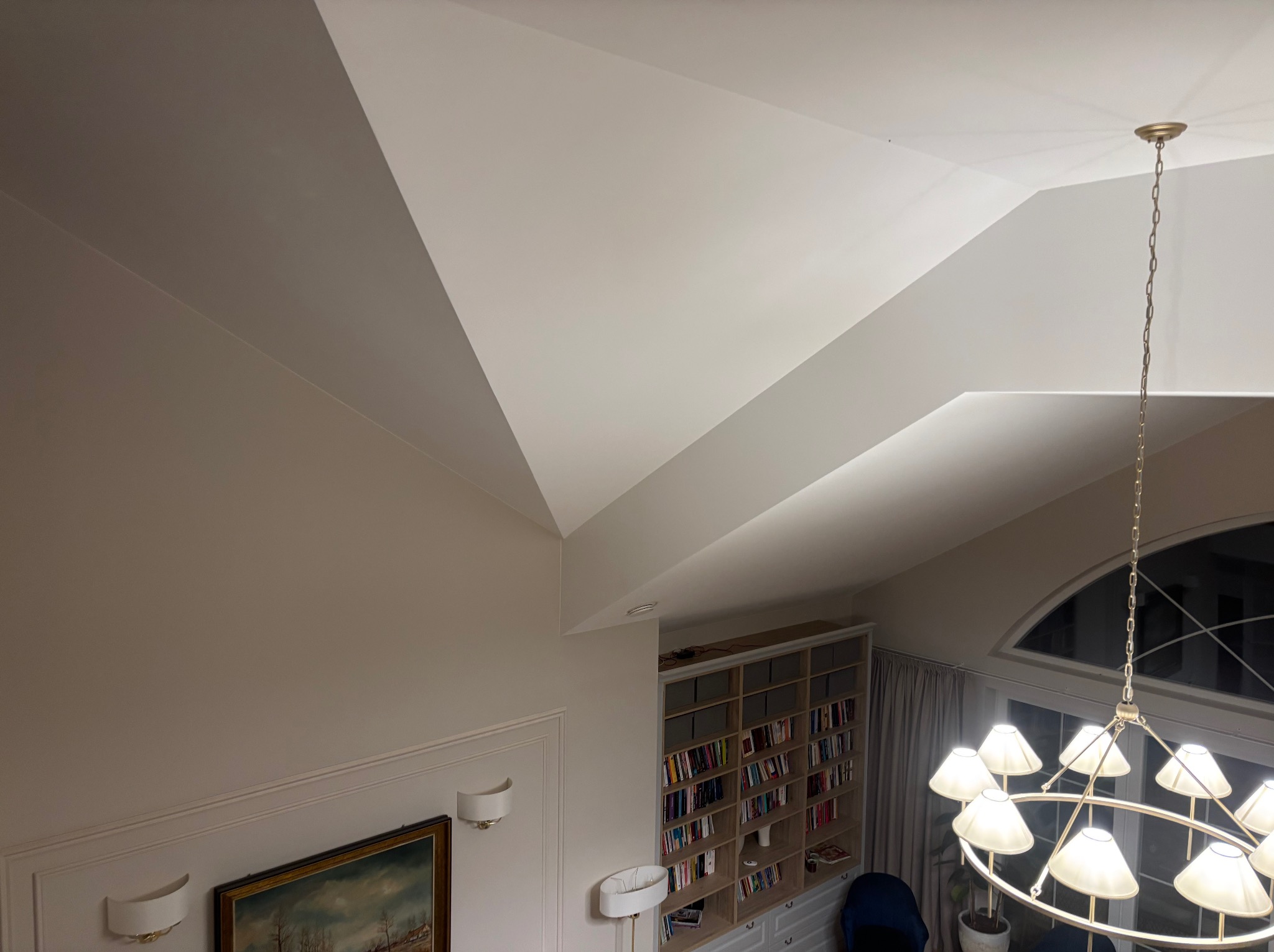 Jasne wnętrze z wysokim skośnym sufitem, oświetlone ozdobnym żyrandolem. Widoczna ściana z obrazem i kinkietami, regał z książkami oraz okno z zasłonami.