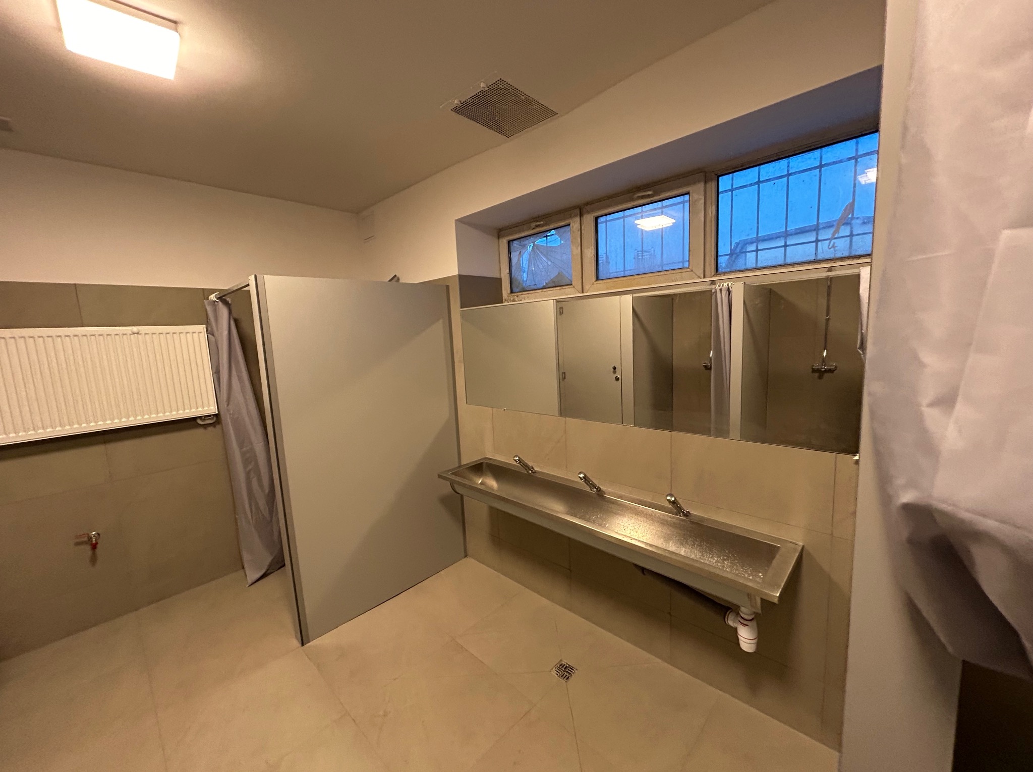 Wykończona łazienka publiczna w Ciechanowie z kabinami prysznicowymi, umywalką ze stali nierdzewnej i grzejnikiem. Minimalistyczny design, neutralna kolorystyka.