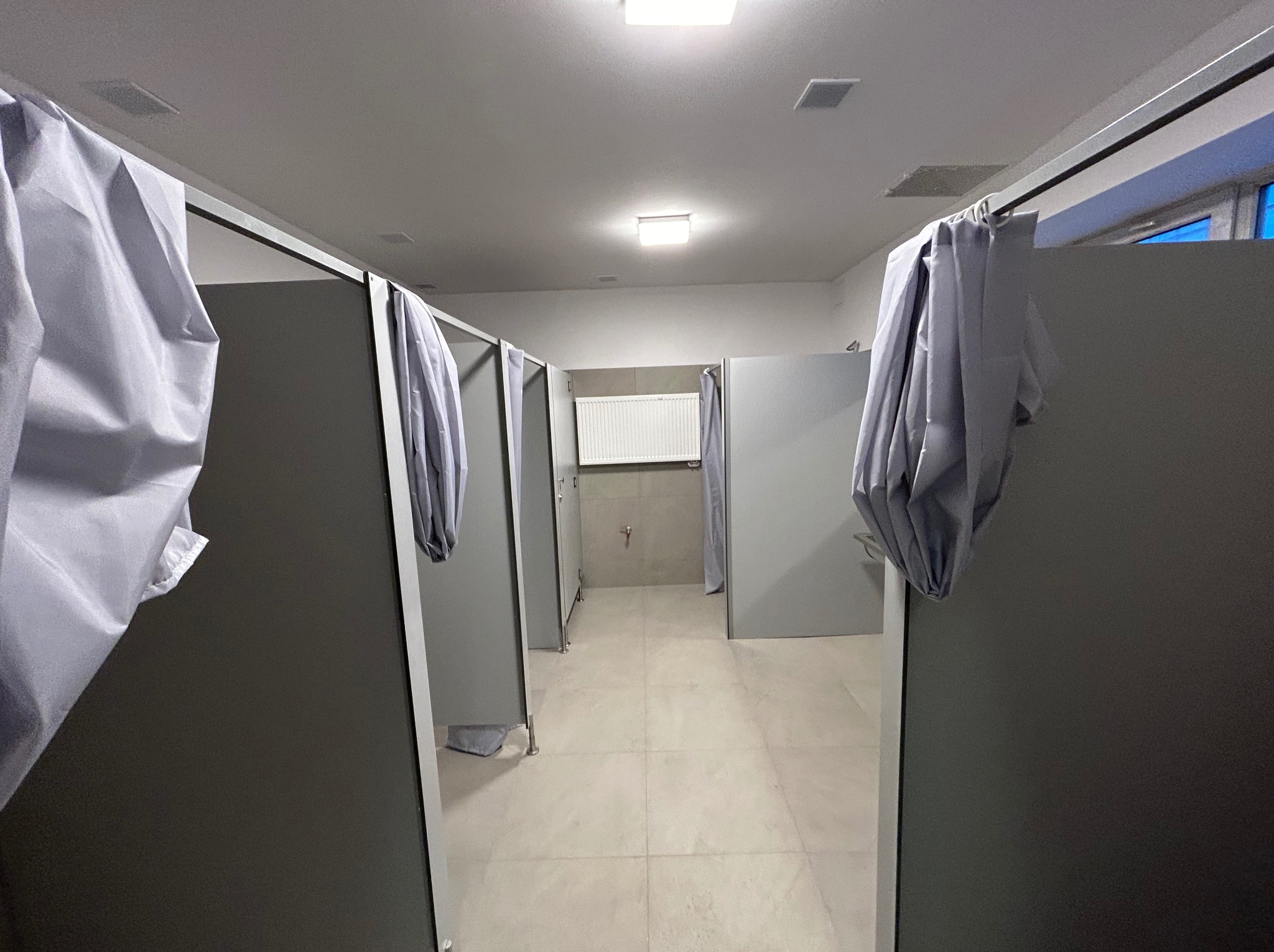 Wnętrze publicznej toalety z kabinami prysznicowymi oddzielonymi szarymi ściankami i zasłonami. Widoczny grzejnik i jasna podłoga z płytek. Minimalistyczny design.