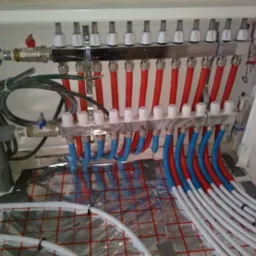 Rozdzielacz ogrzewania podłogowego z czerwonymi i niebieskimi rurami, widoczne złącza i zawory, instalacja w trakcie montażu na folii grzewczej z siatką.