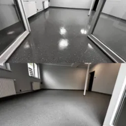 Szara posadzka żywiczna z delikatnymi, ciemnymi inkluzjami w jasnym, pustym pomieszczeniu z białymi grzejnikami i oknami, widoczna przez otwarte, metalowe drzwi.