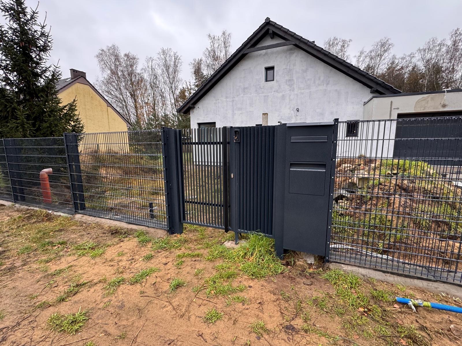 Nowoczesna, grafitowa brama wjazdowa z furtką i skrzynką na listy, wkomponowana w ogrodzenie z siatki na tle domu z szarą elewacją.