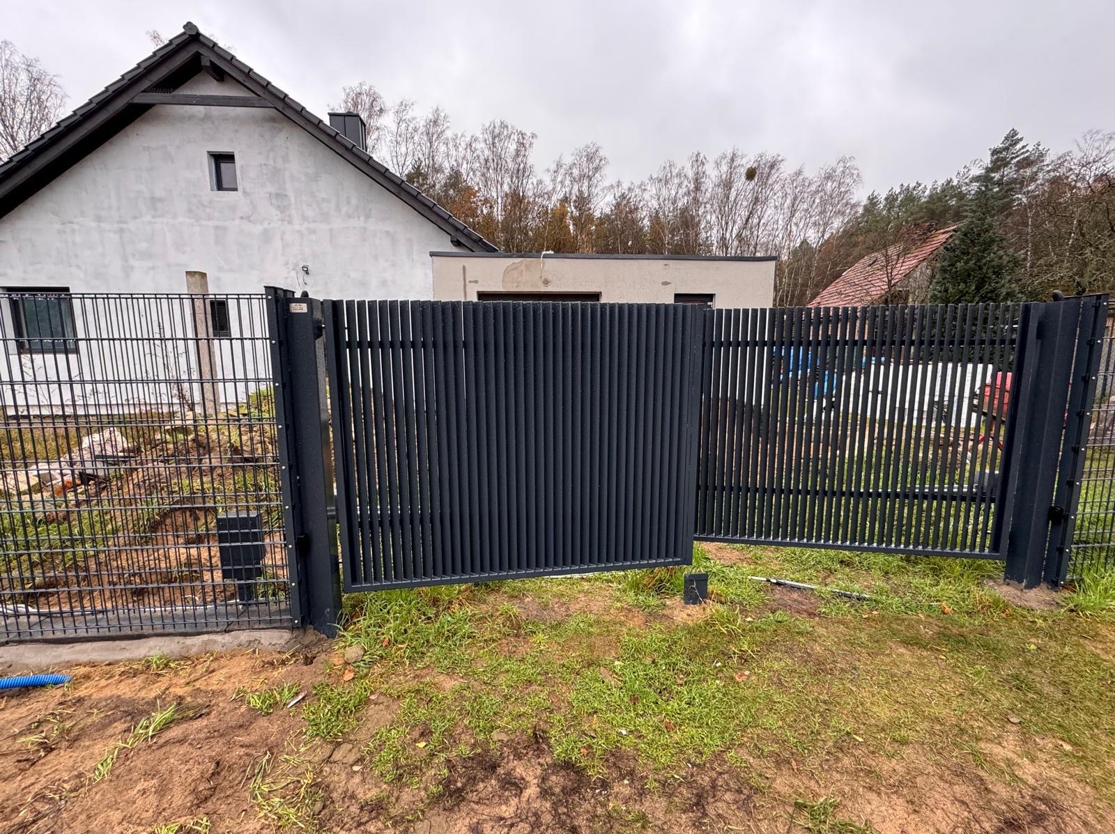 Nowoczesna, przesuwna brama wjazdowa z pionowymi profilami, częściowo otwarta, na tle domu z białym tynkiem i zielenią trawnika.
