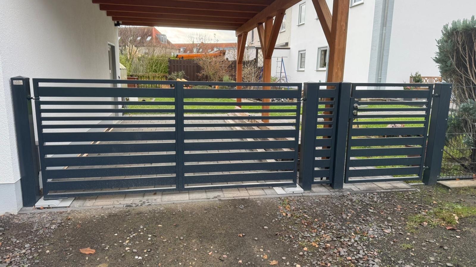 Nowoczesna, pozioma brama wjazdowa z szarego metalu, widoczna od strony podjazdu z kostki brukowej i żwiru, z zadaszeniem wspartym na drewnianych belkach w tle.