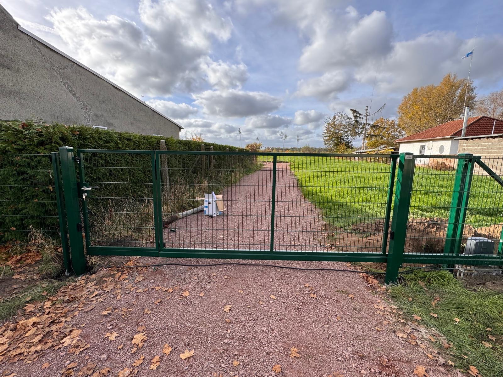 Zielona brama wjazdowa z siatki, widok z zewnątrz posesji, z widoczną skrzynką z akcesoriami montażowymi i kablem zasilającym, na tle drogi z czerwonego żwiru i zielonego trawnika.