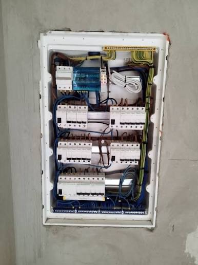 Rozdzielnica elektryczna w białej obudowie, widoczne zabezpieczenia, przewody i listwy zaciskowe. Ściana w surowym stanie, szara.