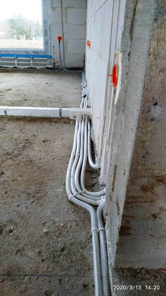 Szare, karbowane rury elektryczne prowadzone wzdłuż surowej ściany z betonu komórkowego, z widocznymi puszkami instalacyjnymi i fragmentem okna w tle.