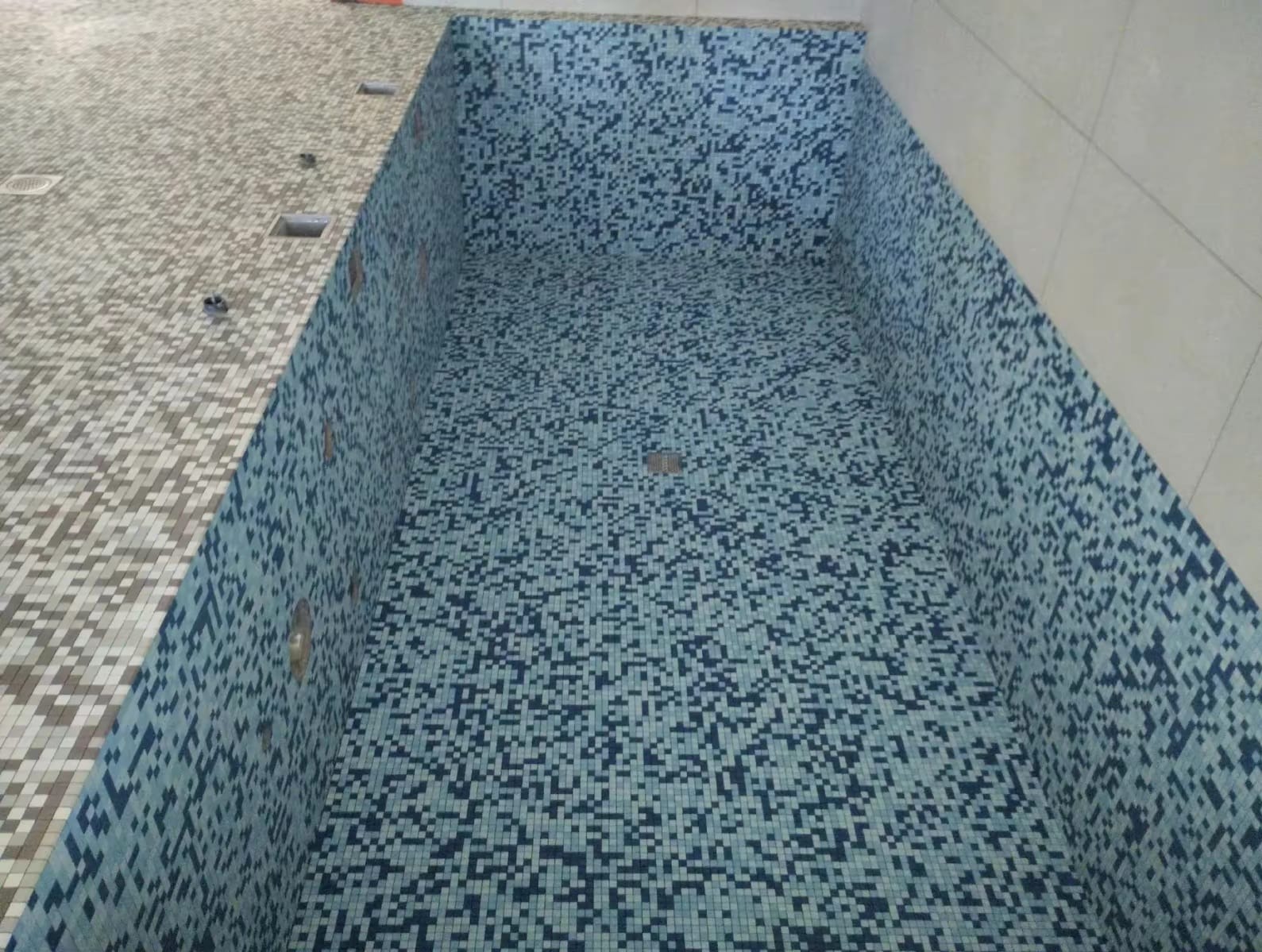 Wnętrze basenu w trakcie wykańczania, wyłożone mozaiką w odcieniach błękitu i granatu, z widocznymi otworami na dysze wodne i odpływem w dnie.