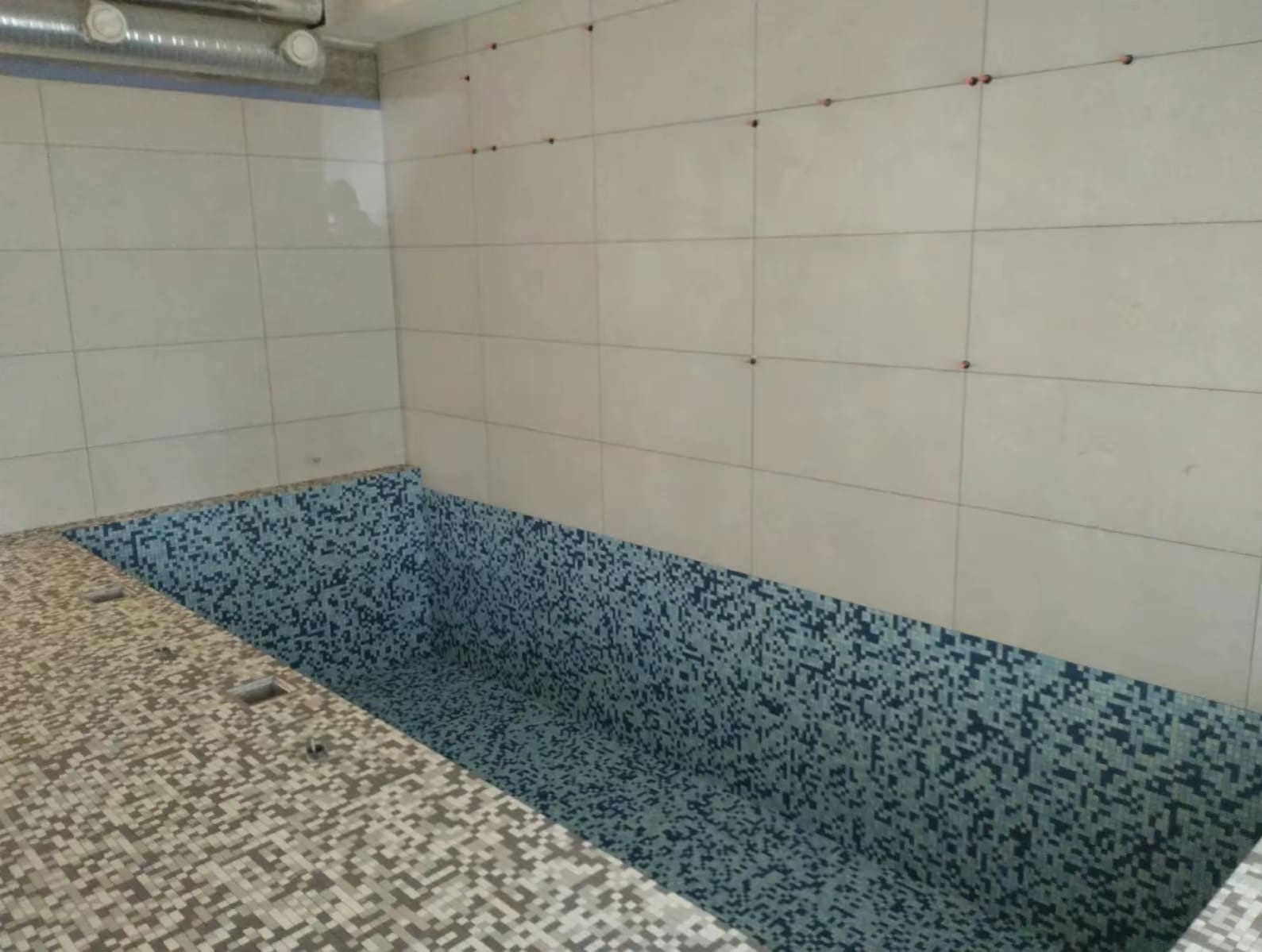 Wnętrze basenu w trakcie wykańczania, z mozaiką w odcieniach niebieskiego i szarego, oraz jasnymi płytkami na ścianach z widocznymi elementami poziomowania.