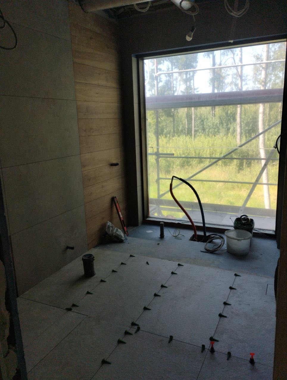 Wnętrze łazienki w trakcie układania szarych płytek podłogowych z użyciem krzyżyków dystansowych, widoczne narzędzia i materiały budowlane przy oknie z widokiem na las.