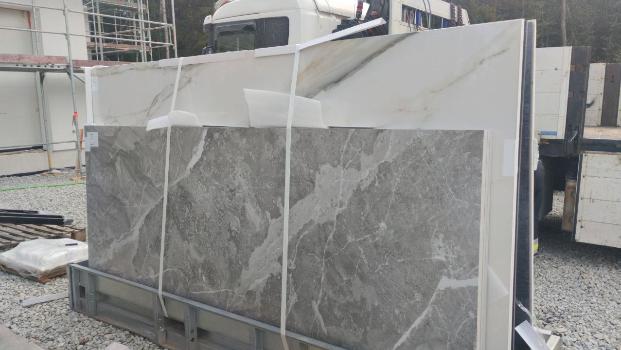 Płyty wielkoformatowe z marmurowym wzorem, szare i białe, zabezpieczone taśmą, na metalowej palecie transportowej, widoczna ciężarówka i rusztowanie w tle.