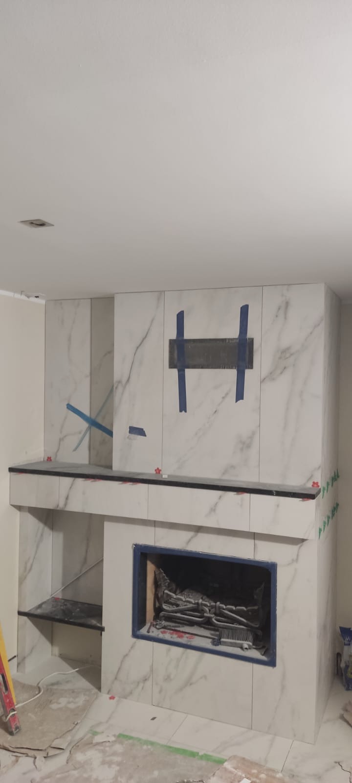 Montaż marmurowych płytek wokół kominka, widoczne niebieskie taśmy maskujące, poziomica i plastikowe krzyżyki dystansowe, podłoga z resztkami zaprawy.