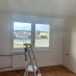 Denys Karpovych - Pokój w trakcie odświeżania: białe ściany, okno z widokiem na miasto, drabina z narzędziami. Częściowo pomalowana ściana i brązowa podłoga.