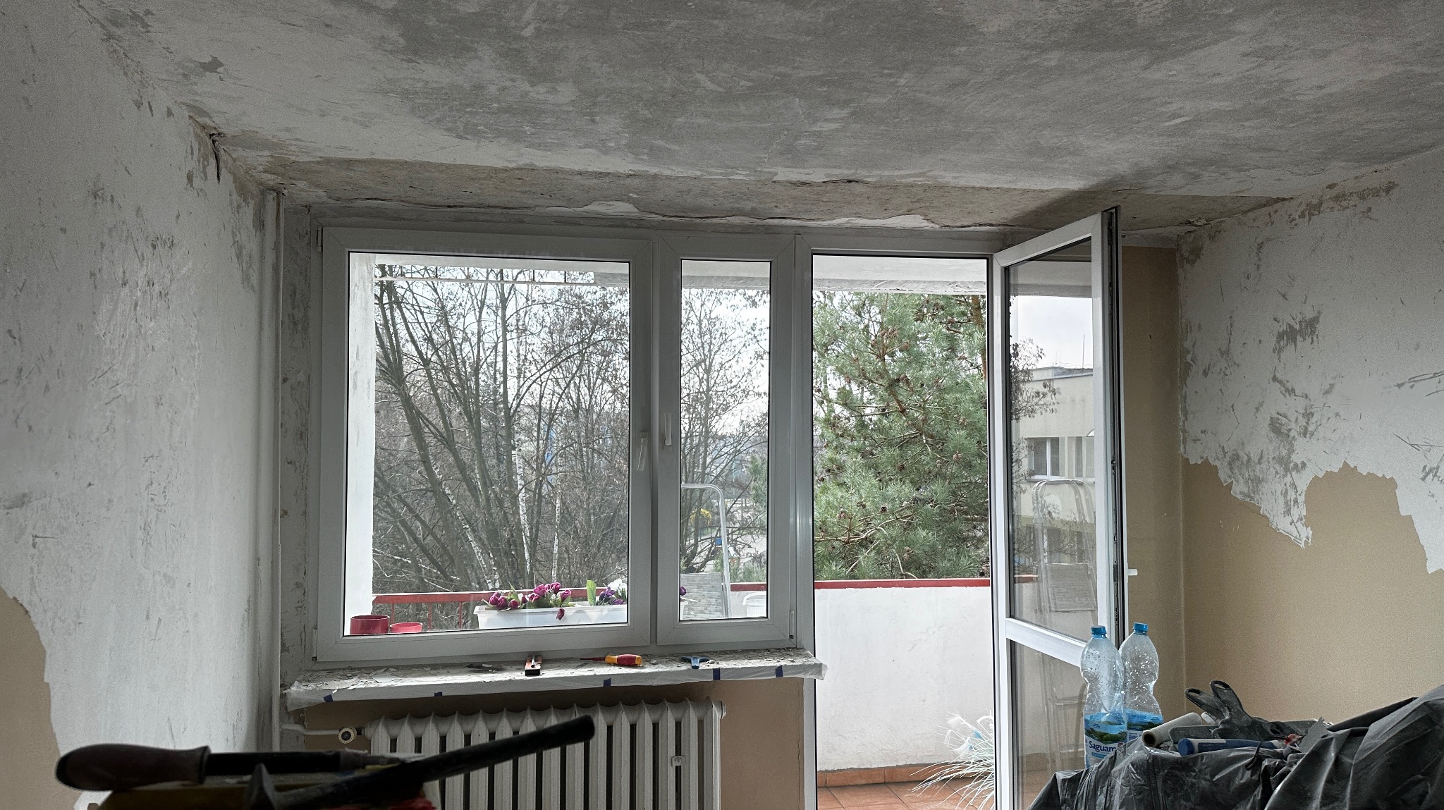 Pokój w trakcie przygotowań do remontu: ściany z resztkami farby, okno z widokiem na drzewa, narzędzia i butelki na parapecie, otwarte drzwi balkonowe.