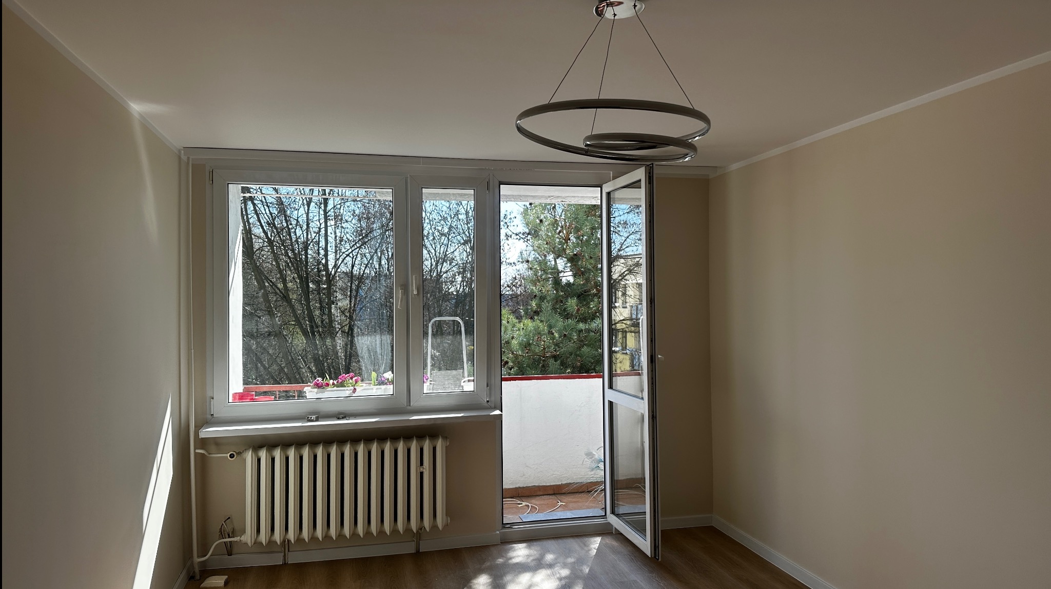 Jasne wnętrze pokoju po malowaniu ścian na beżowo z widokiem na balkon i nowoczesną lampą sufitową.