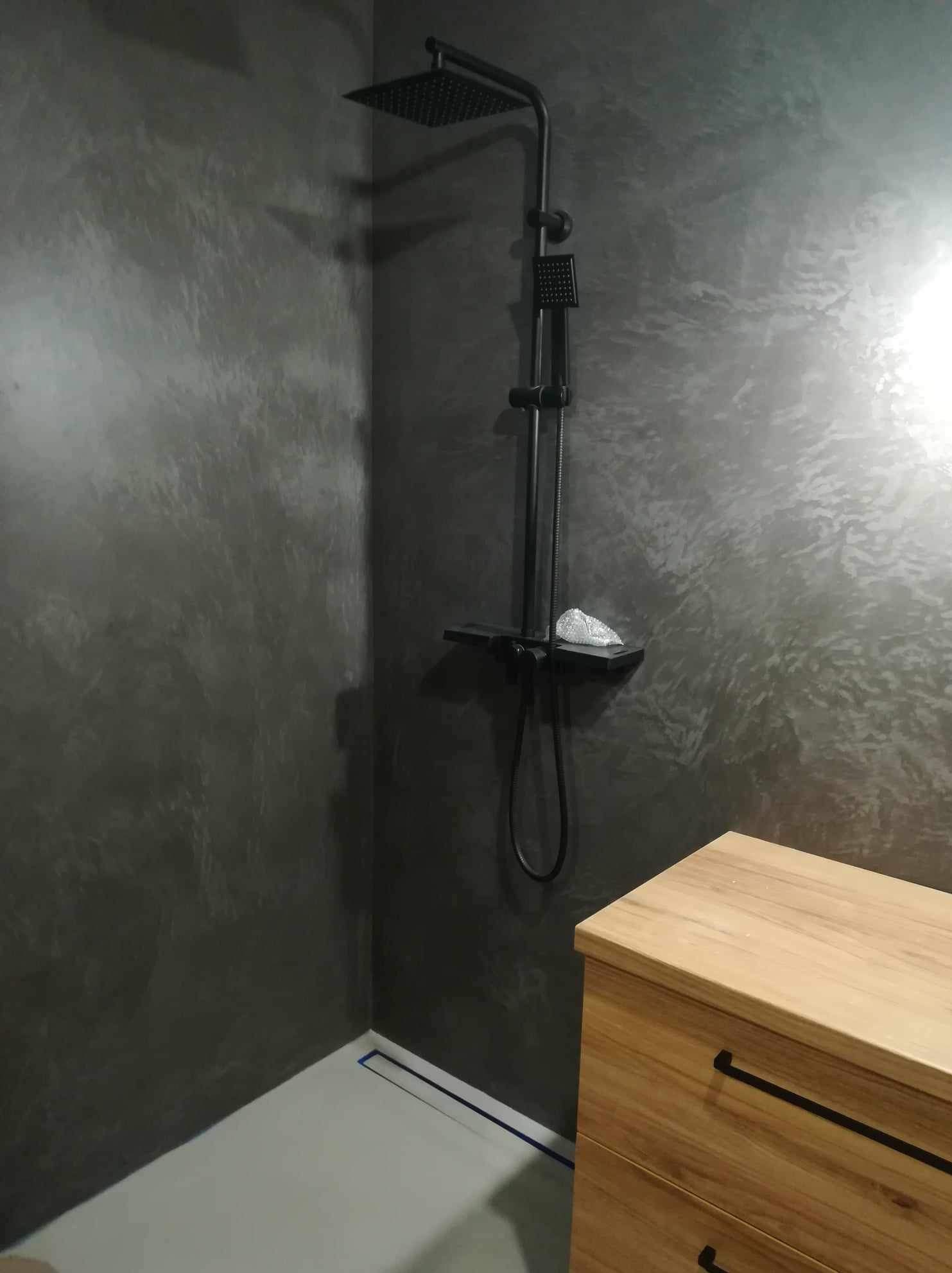 Nowoczesna kabina prysznicowa z czarną armaturą i dekoracyjną szarą ścianą. Widoczny odpływ liniowy i drewniana szafka łazienkowa.