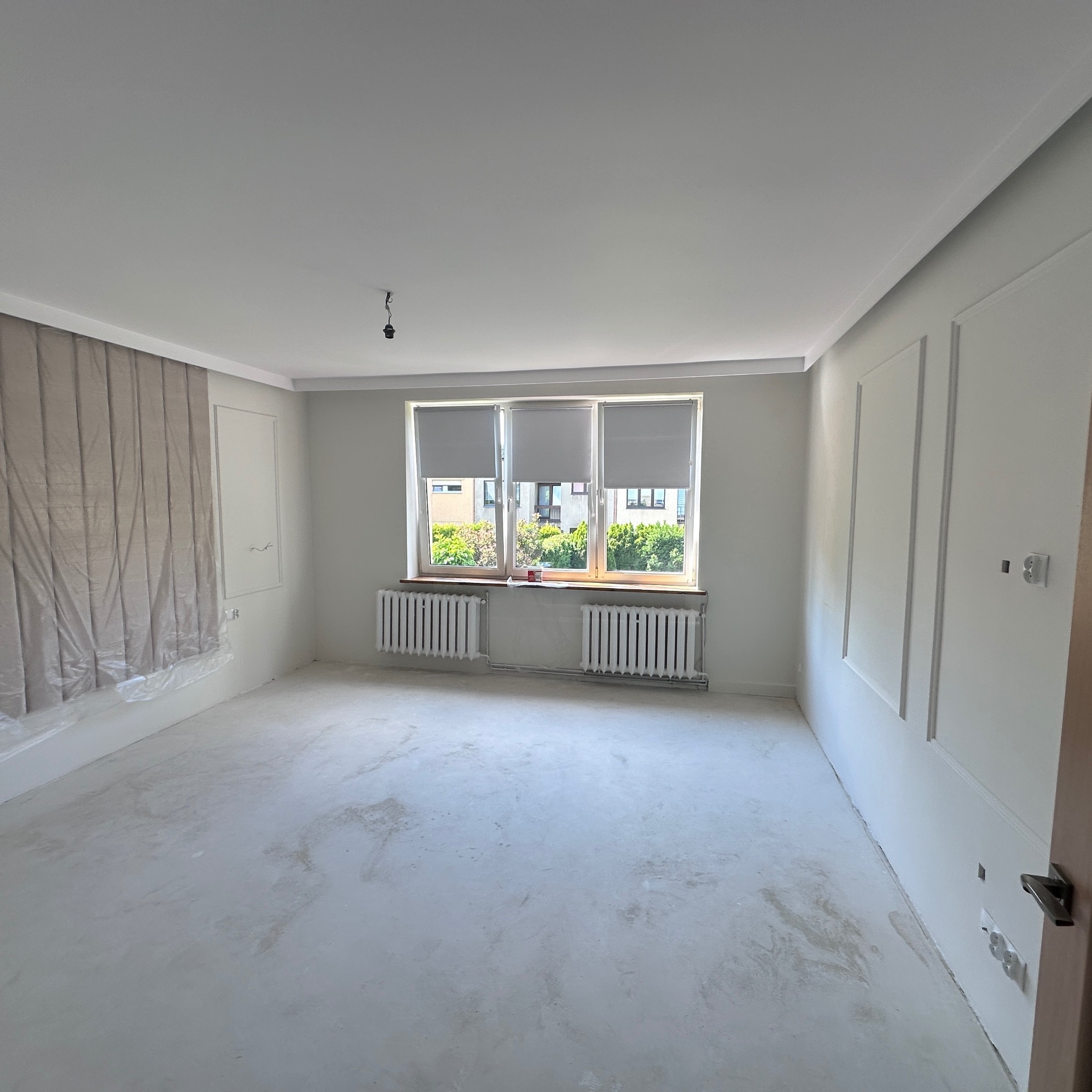 Jasne wnętrze po gładziowaniu ścian i podłogi, okno z roletami, grzejniki, dekoracyjne listwy ścienne. Folia ochronna zabezpiecza fragment ściany.