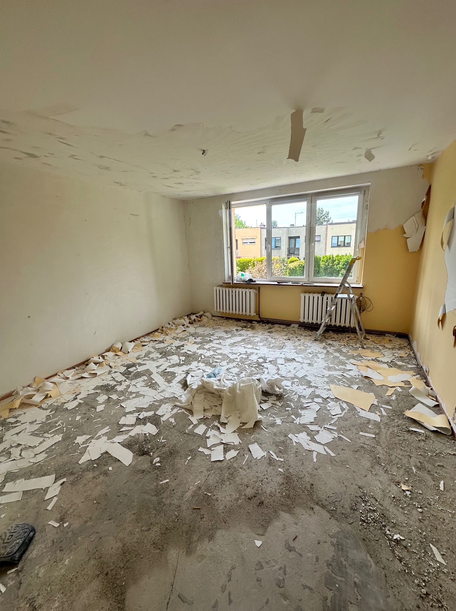 Pokój w trakcie usuwania starych tapet, widoczne resztki tapet na podłodze i ścianach. Drabina oparta o ścianę przy oknie. Przygotowanie do remontu.