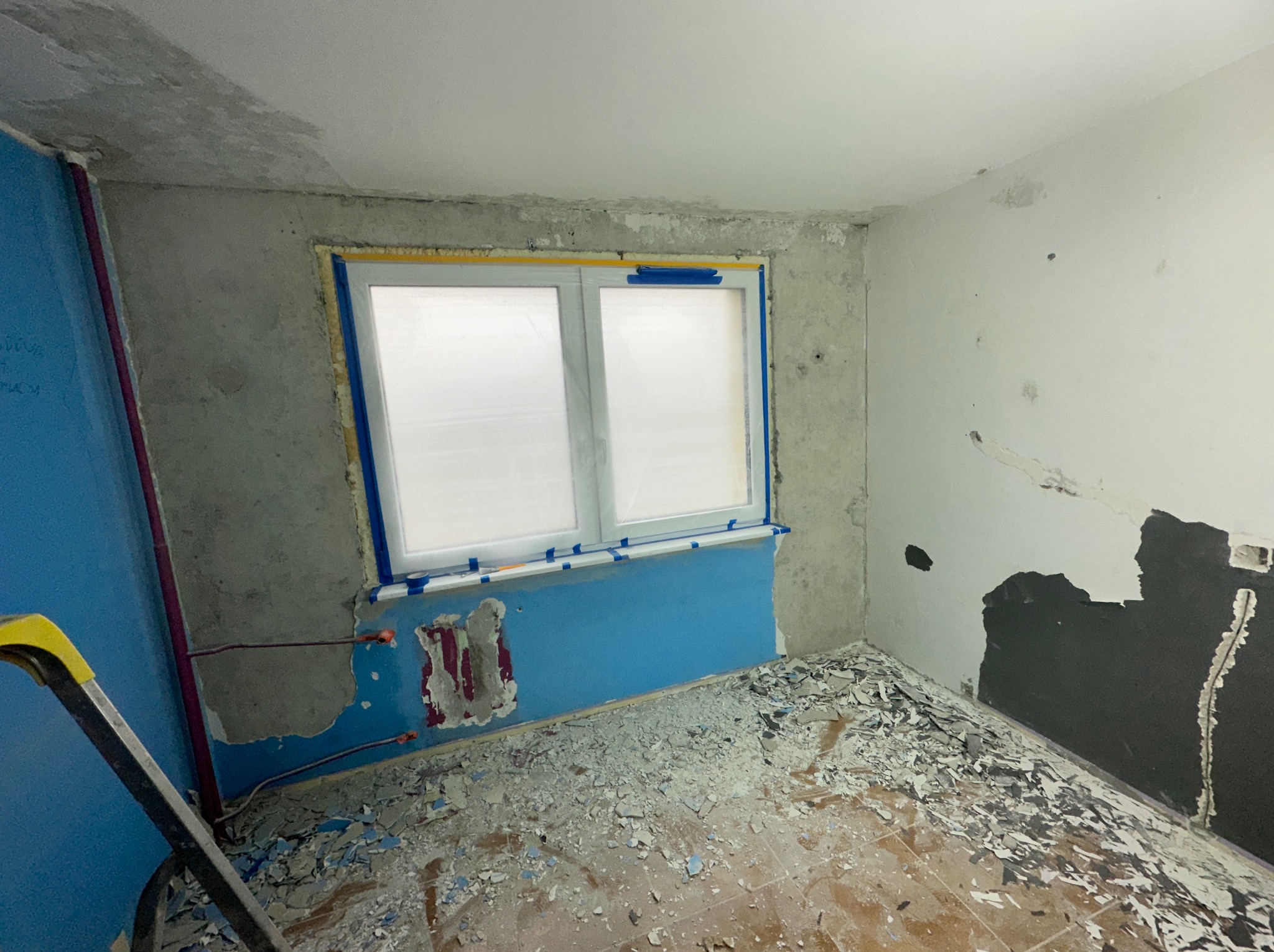 Pokój w trakcie przygotowań do remontu: zabezpieczone okno folią i taśmą, ściany częściowo obdrapane, podłoga pokryta gruzem, widoczna drabina malarska.