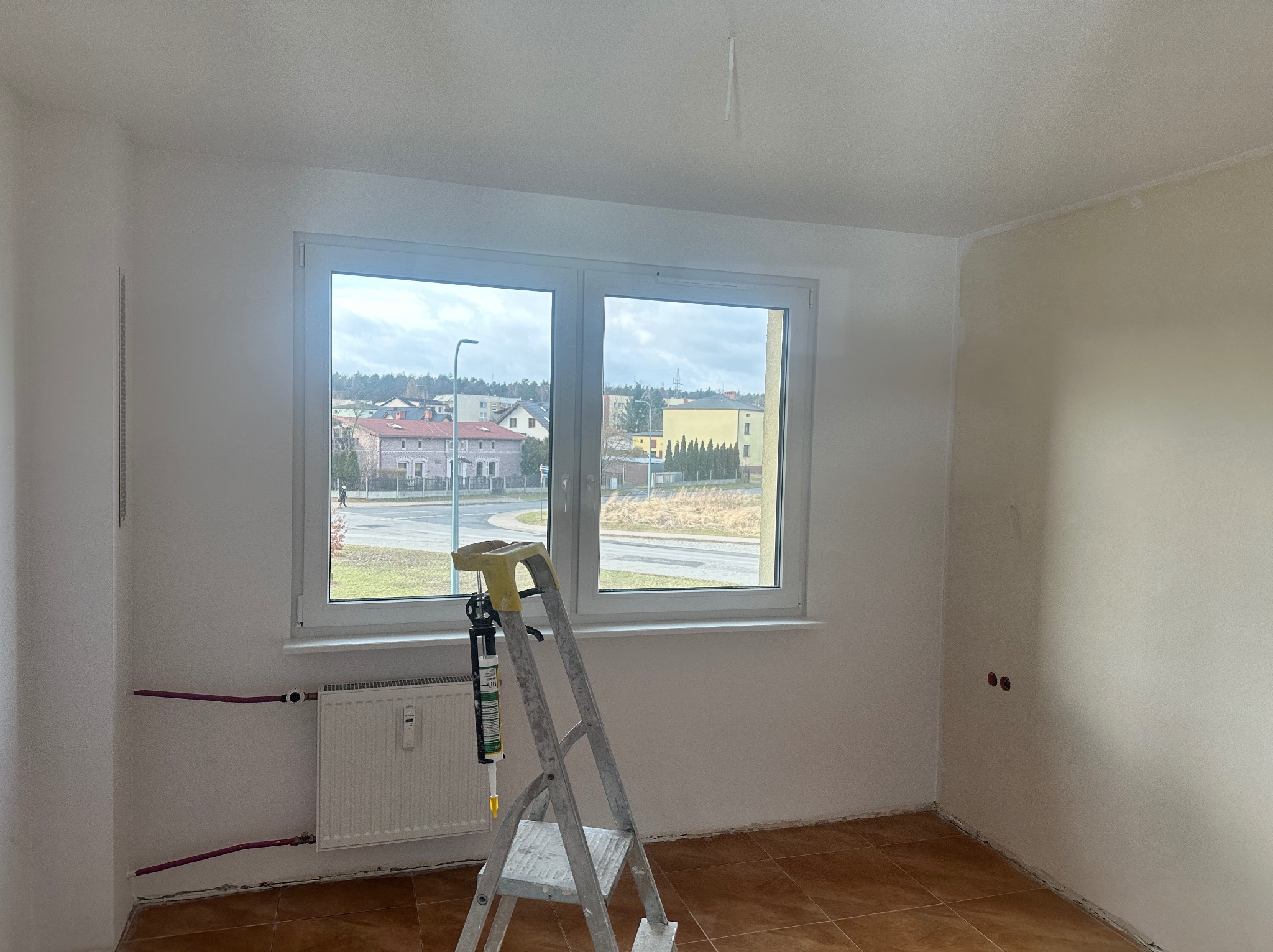Pokój w trakcie odświeżania: białe ściany, okno z widokiem na miasto, drabina z narzędziami. Częściowo pomalowana ściana i brązowa podłoga.