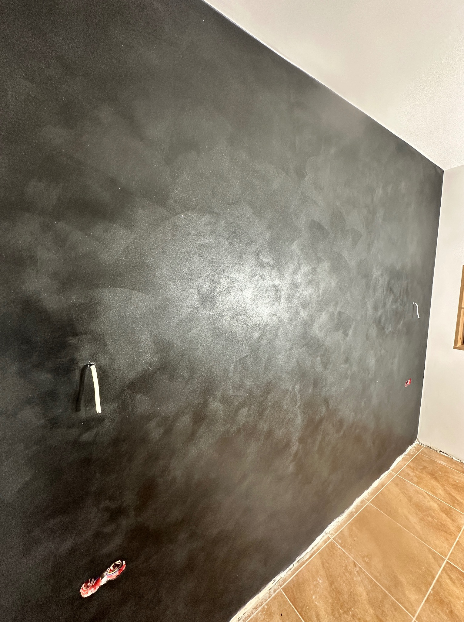 Ciemna, błyszcząca ściana z efektem dekoracyjnym w wykończonym wnętrzu. Widoczne wystające kable i fragment podłogi z płytek. Nietypowa faktura ściany.