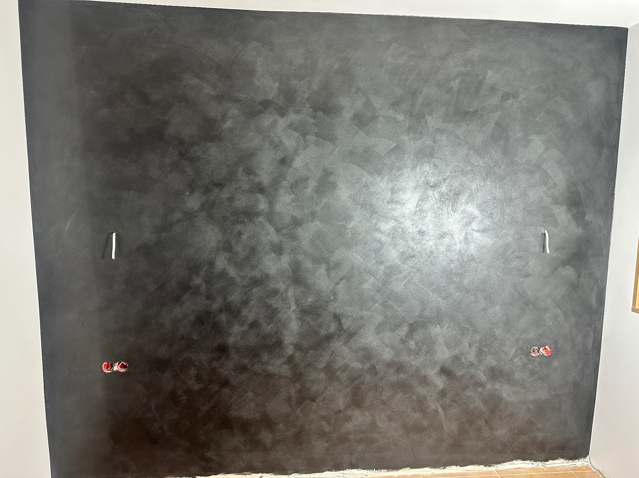 Ściana pomalowana na ciemny, metaliczny kolor z widoczną fakturą. Widoczne puszki elektryczne i fragment podłogi z płytek.